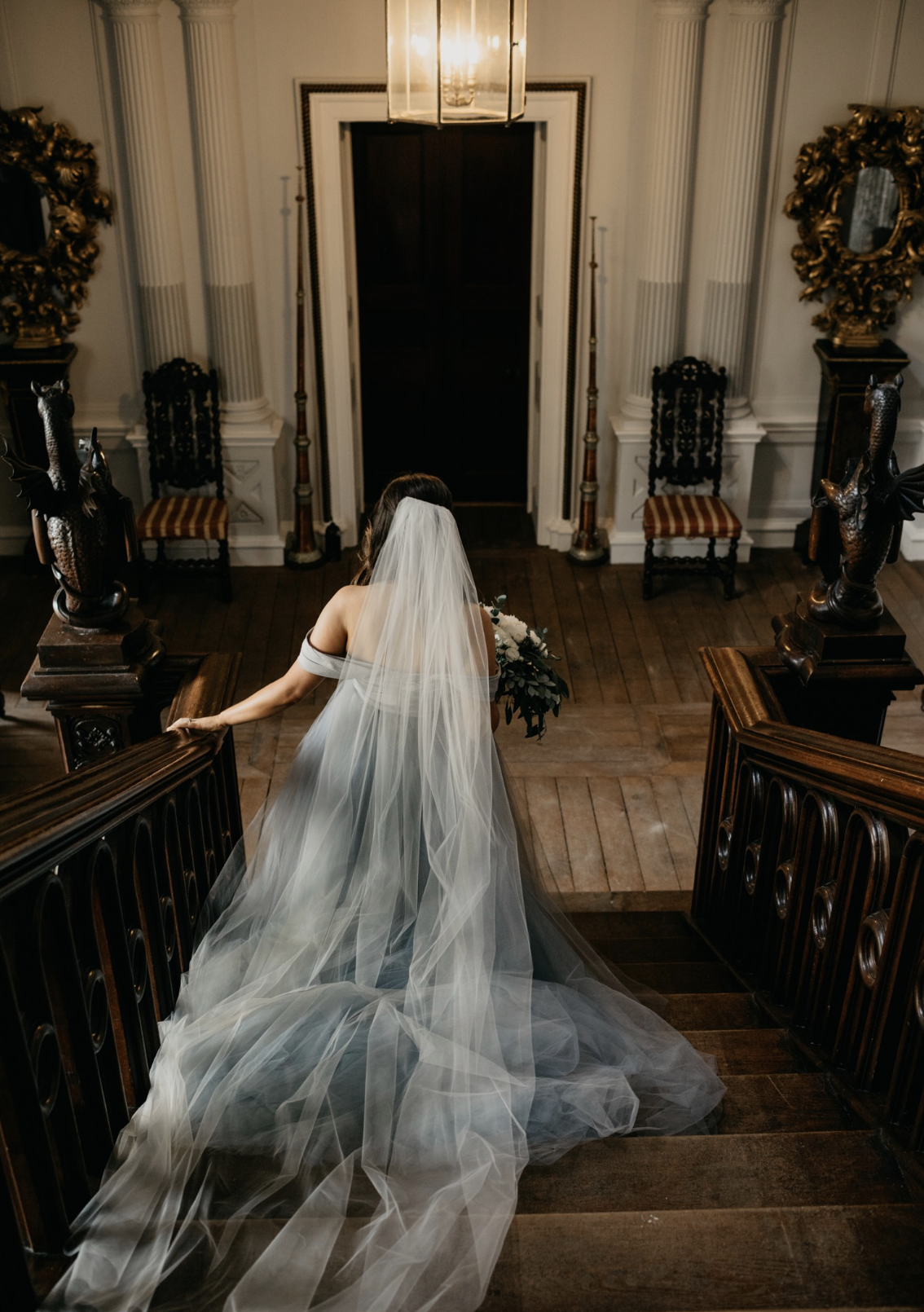 Bridal bride in a flowing wedding gown and veil walking down carved wooden staircase holding bouquet in elegant mansion.