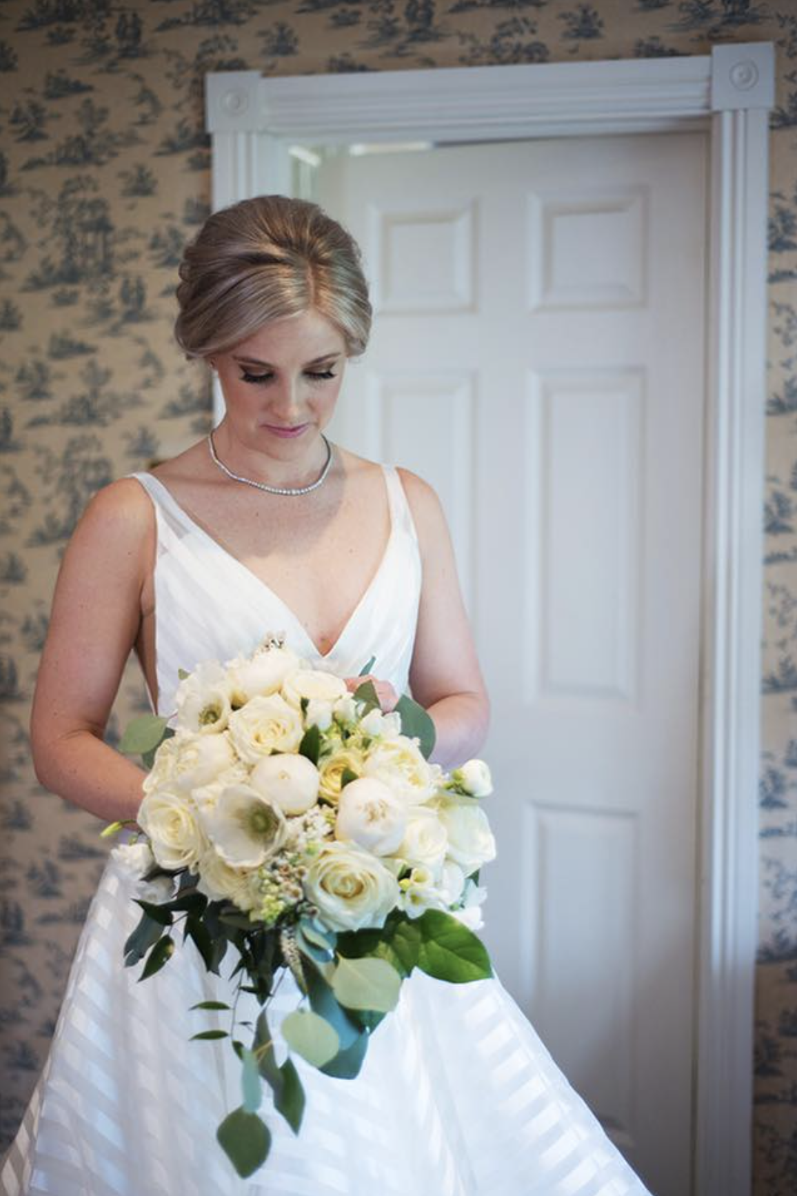 A bride is standing indoors holding a bouquet of white flowers, wearing a white dress with thin straps, and looking down towards her bouquet.