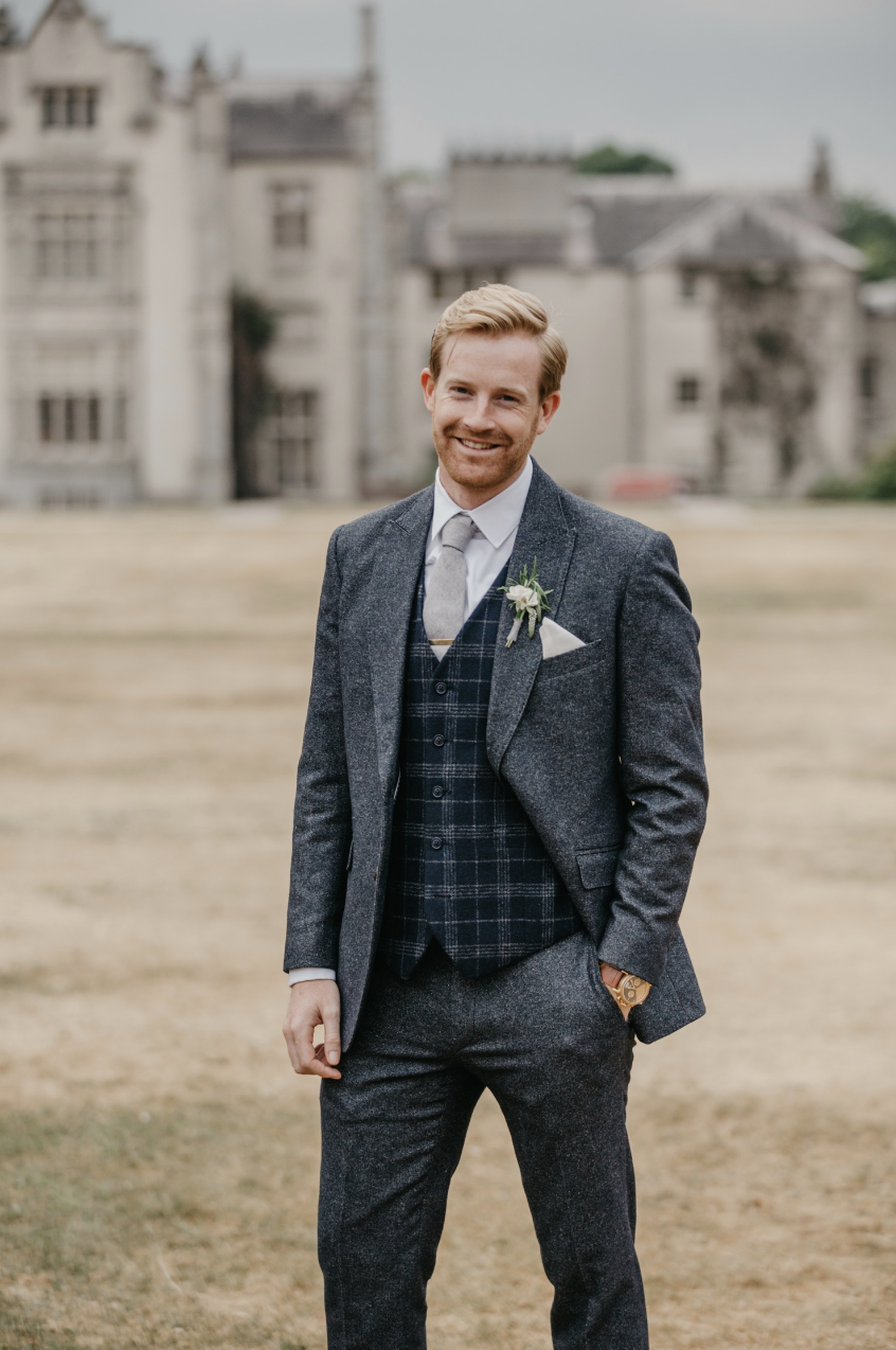 A smiling man in a gray suit with a plaid vest and tie, standing outdoors on a grassy area with a large, historic building in the background.