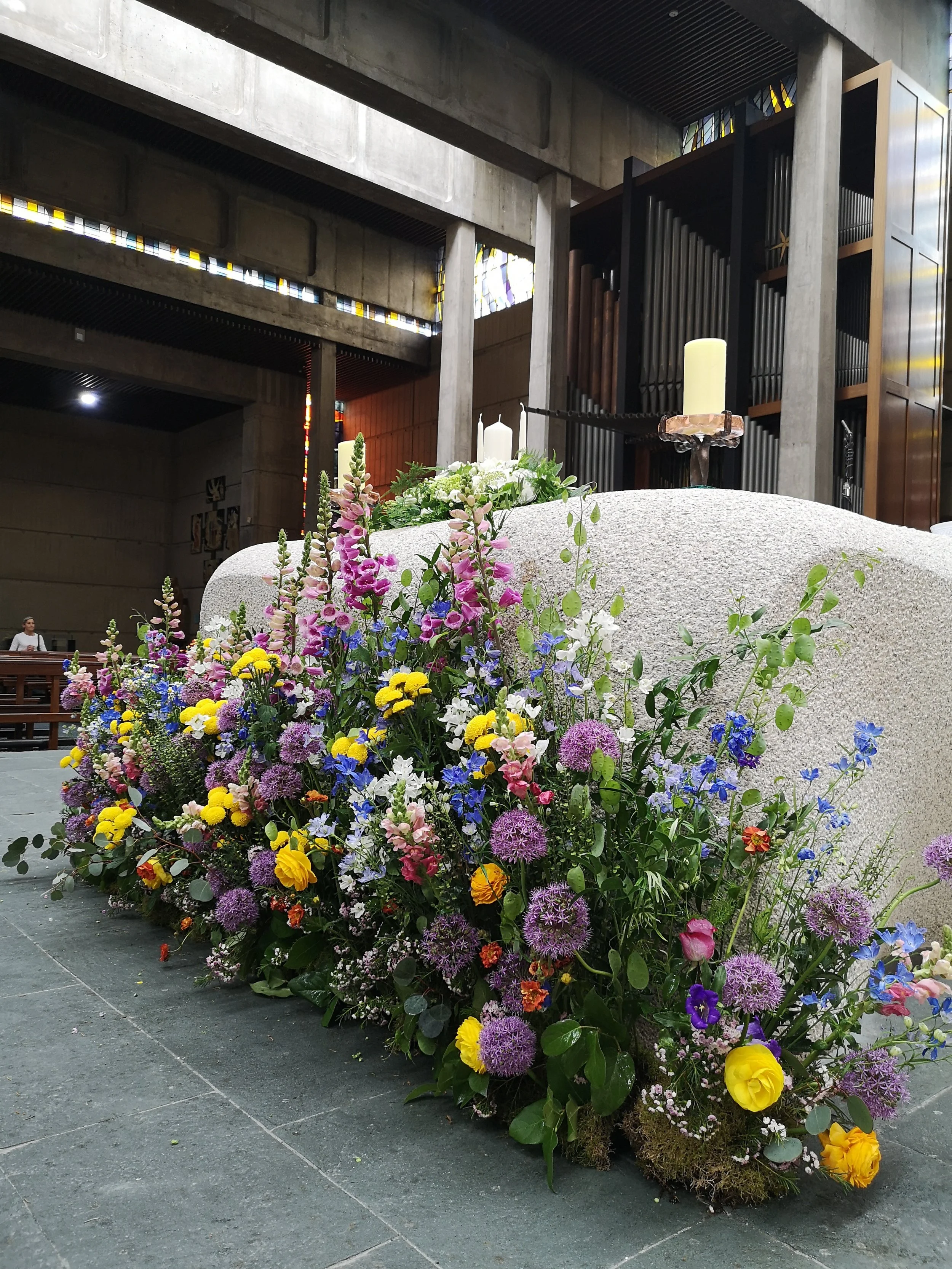 Colorful flower arrangement with yellow, purple, pink, white, and blue flowers placed on the floor in front of a stone altar with candles inside a church.