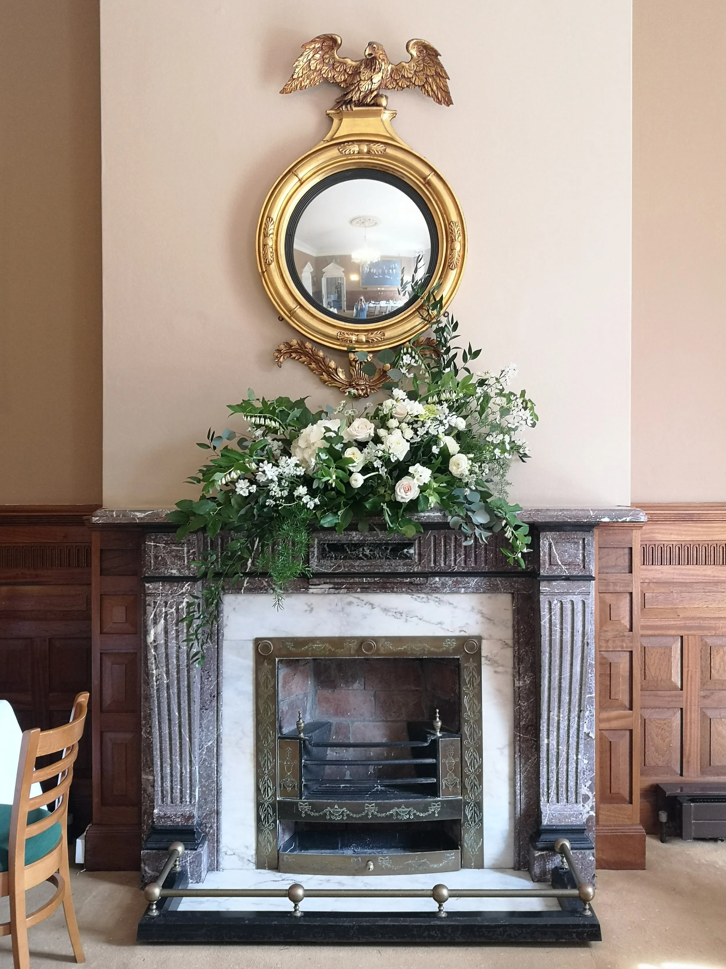 A fireplace with a floral arrangement on the mantel, a circular mirror with a gold frame above it, topped with a decorative eagle. The fireplace is surrounded by wood paneling and grey stonework.