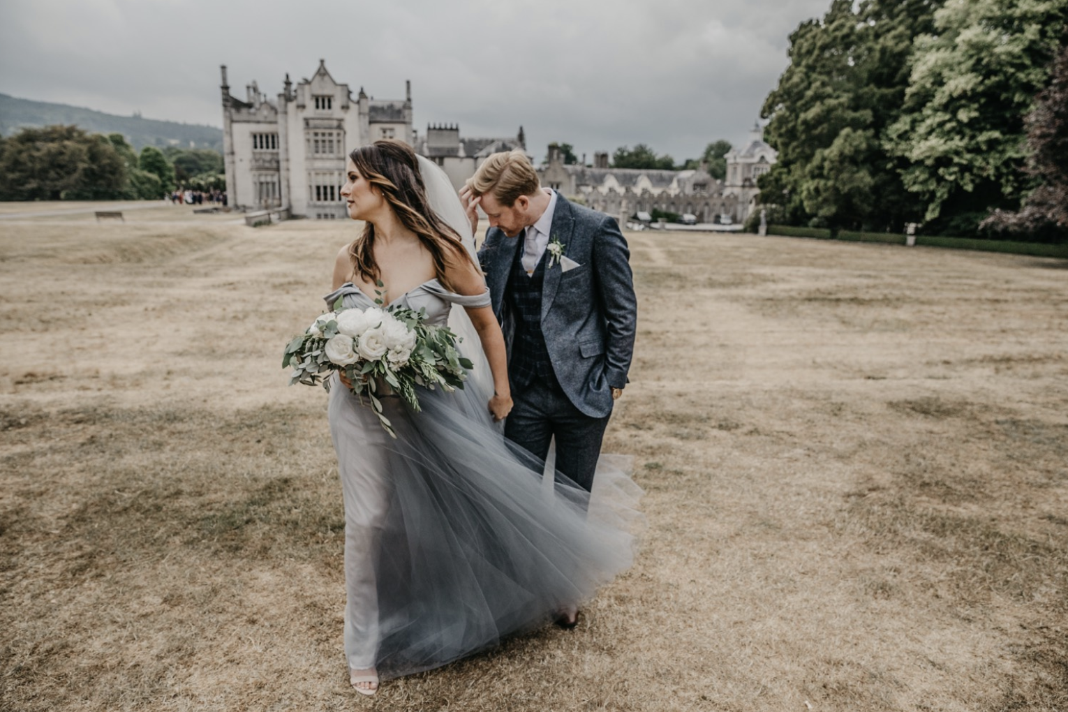 A bride and groom walking on a large, open grassy area outside a historic castle-like building. The bride is holding a bouquet of white flowers and wearing a flowing gray gown, while the groom is dressed in a dark gray suit with a checked vest. The c