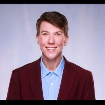 Professional headshot of a person with short brown hair, wearing a maroon blazer and a light blue shirt, smiling against a light blue background.