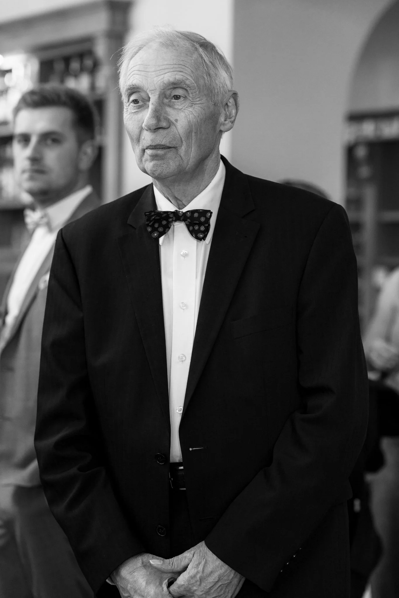 Ein älterer Mann trägt einen schwarzen Anzug mit weiße Hemd und Fliege, steht in einem Raum mit anderen Menschen im Hintergrund.