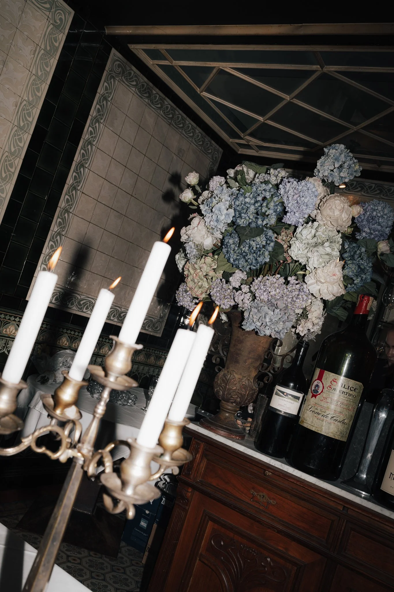 Ein Blumenstrauß aus blauen und weißen Hortensien in einer antiken Vase auf einem Holzschrank, umgeben von Weinflaschen und Kerzen auf einem festlich gedeckten Tisch in einem eleganten Raum.
