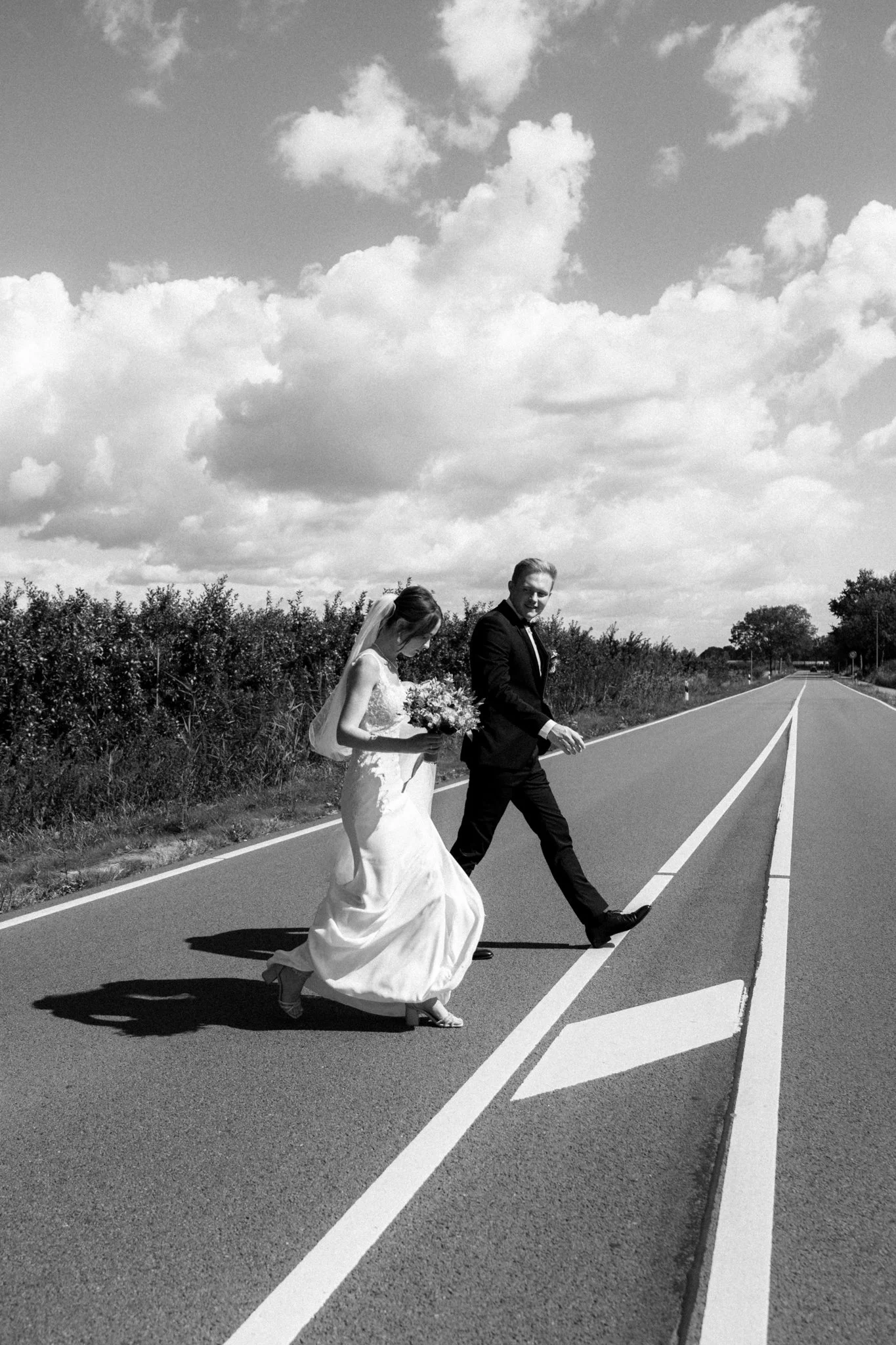 Ein Hochzeitsfoto in Schwarz-Weiß von einem Brautpaar, das auf einer Straße zu Fuß geht. Der Himmel ist mit Wolken bedeckt.