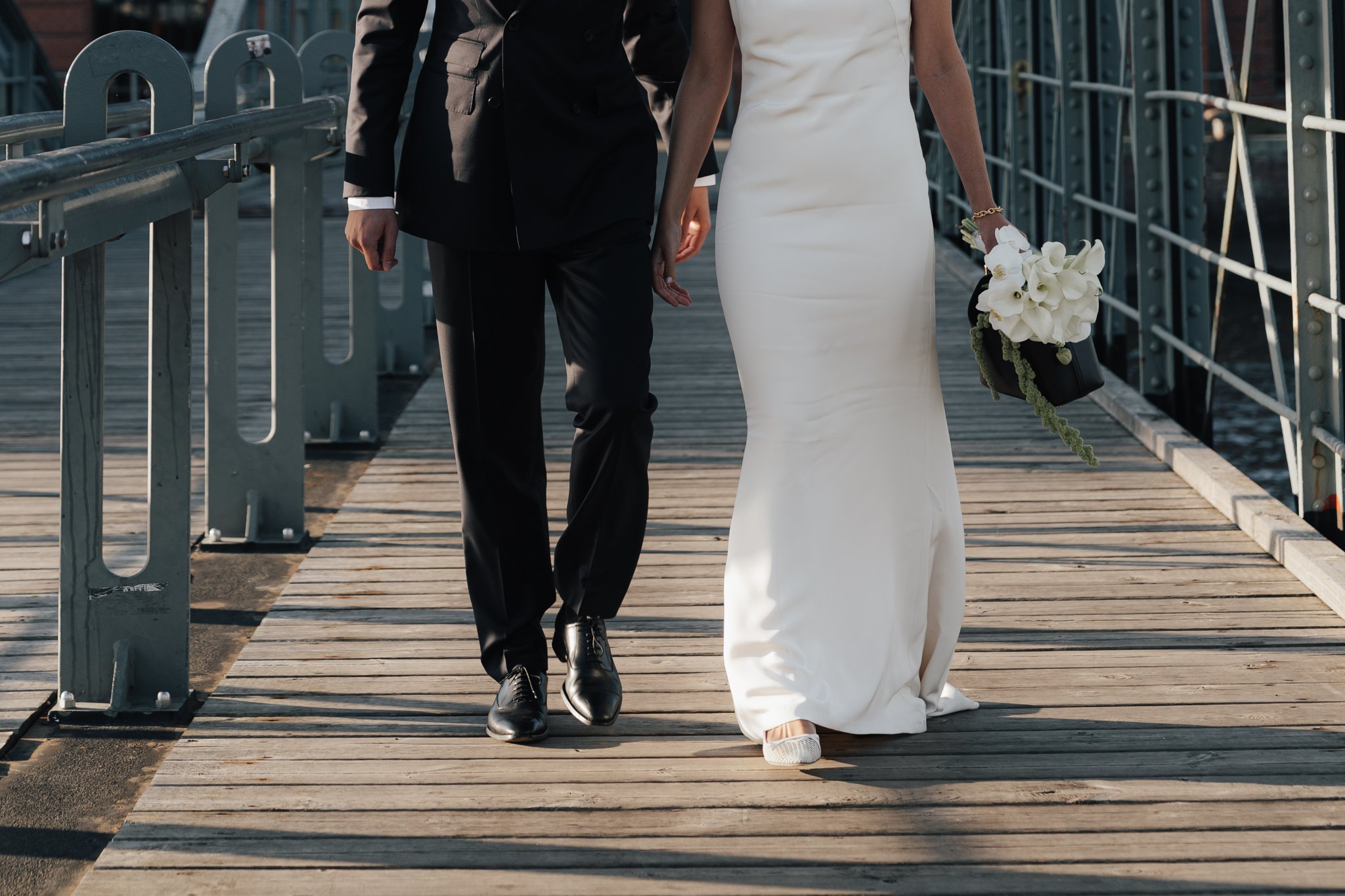 Ein Brautpaar läuft auf einer Holzbrücke, die Frau trägt ein langes weißes Brautkleid und hält einen Blumenstrauß, der Mann trägt einen schwarzen Anzug.