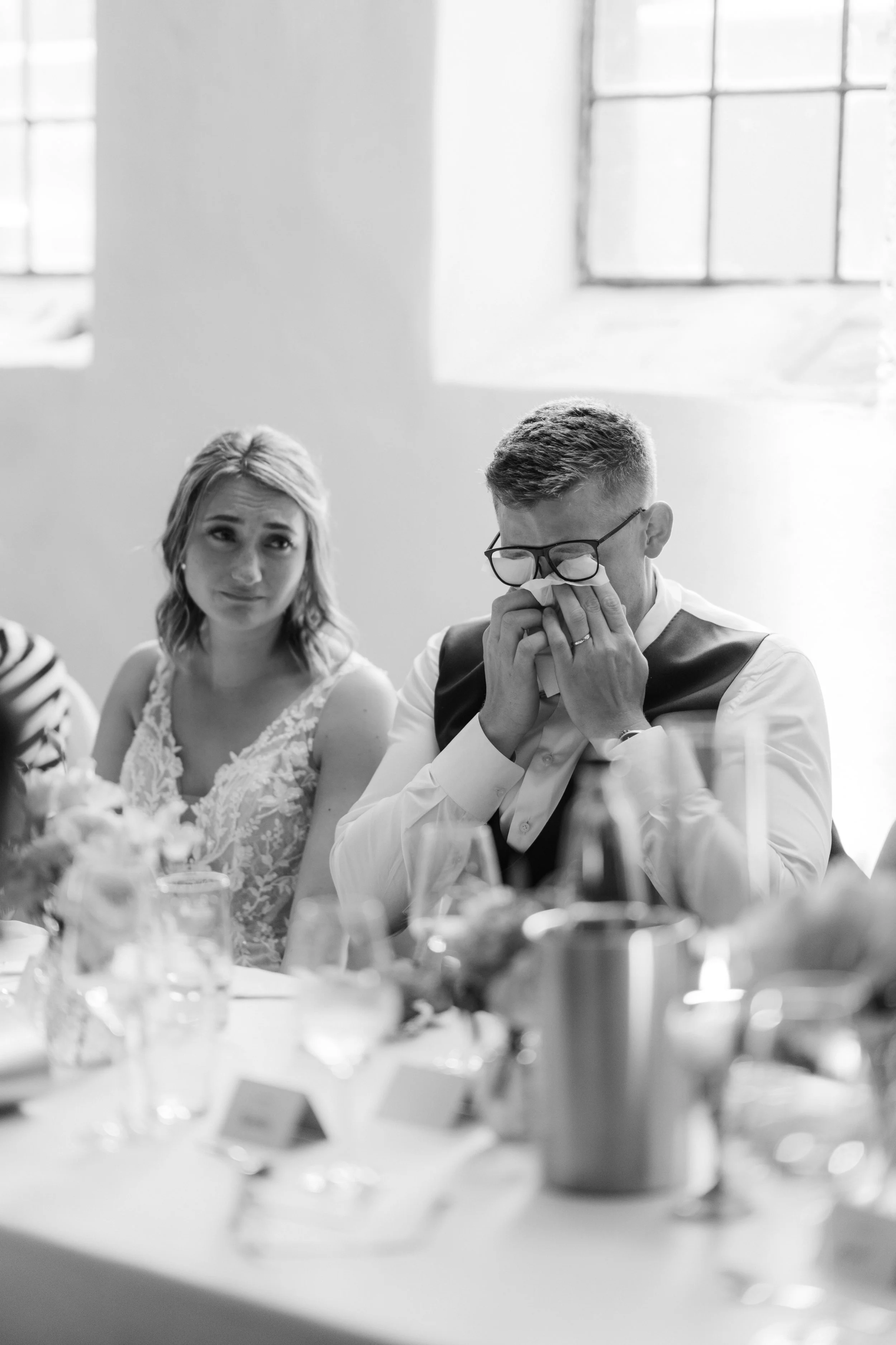Ein emotionaler Moment bei einer Hochzeit, ein Mann weint, während eine Frau neben ihm sitzt, im Hintergrund ein schiefer Blick auf ein Fenster.