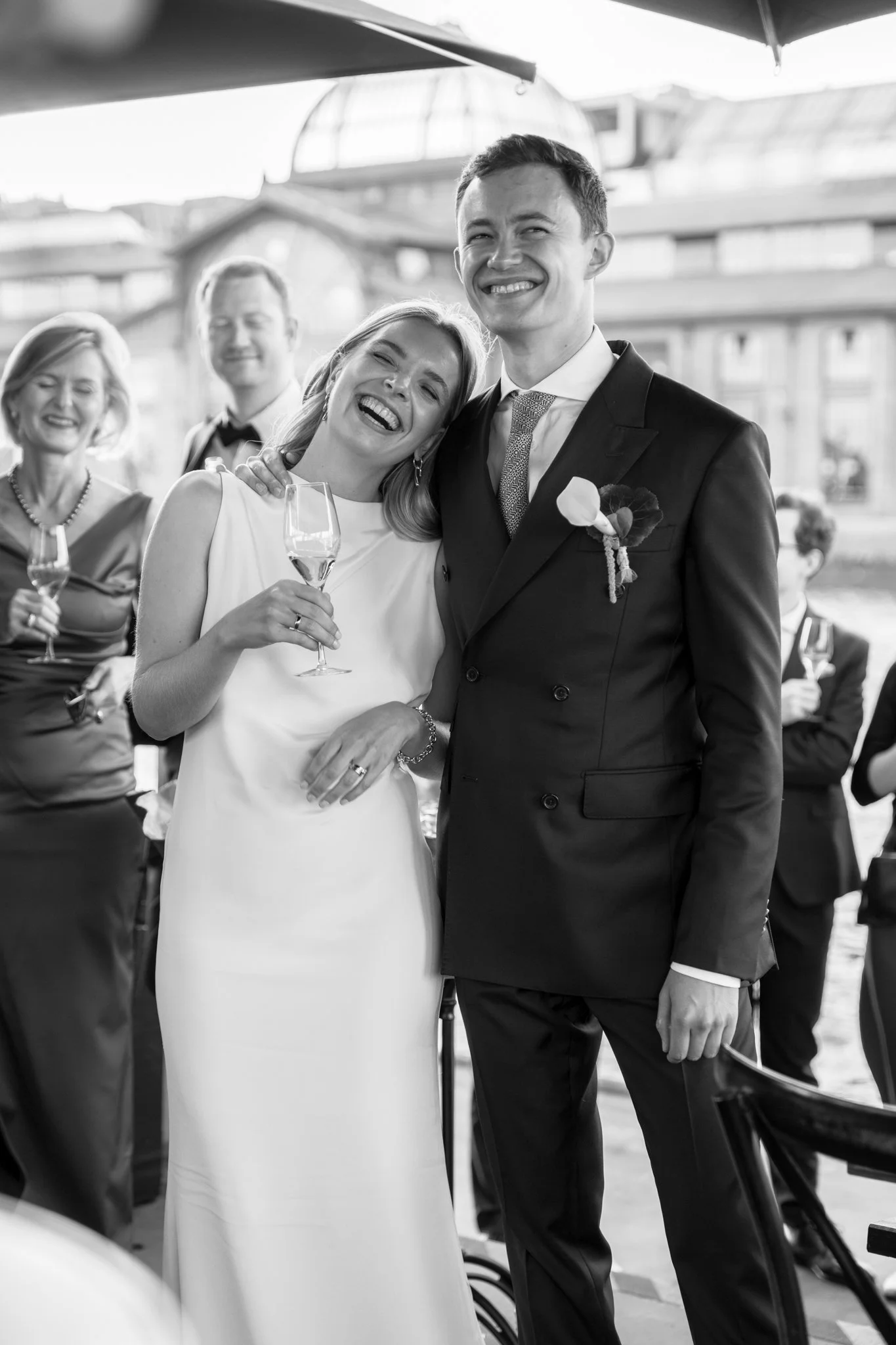 Ein glückliches Brautpaar bei ihrer Hochzeit, lachend und mit Sekt in der Hand, umgeben von Freunden an einem festlichen Anlass, vermutlich im Freien.