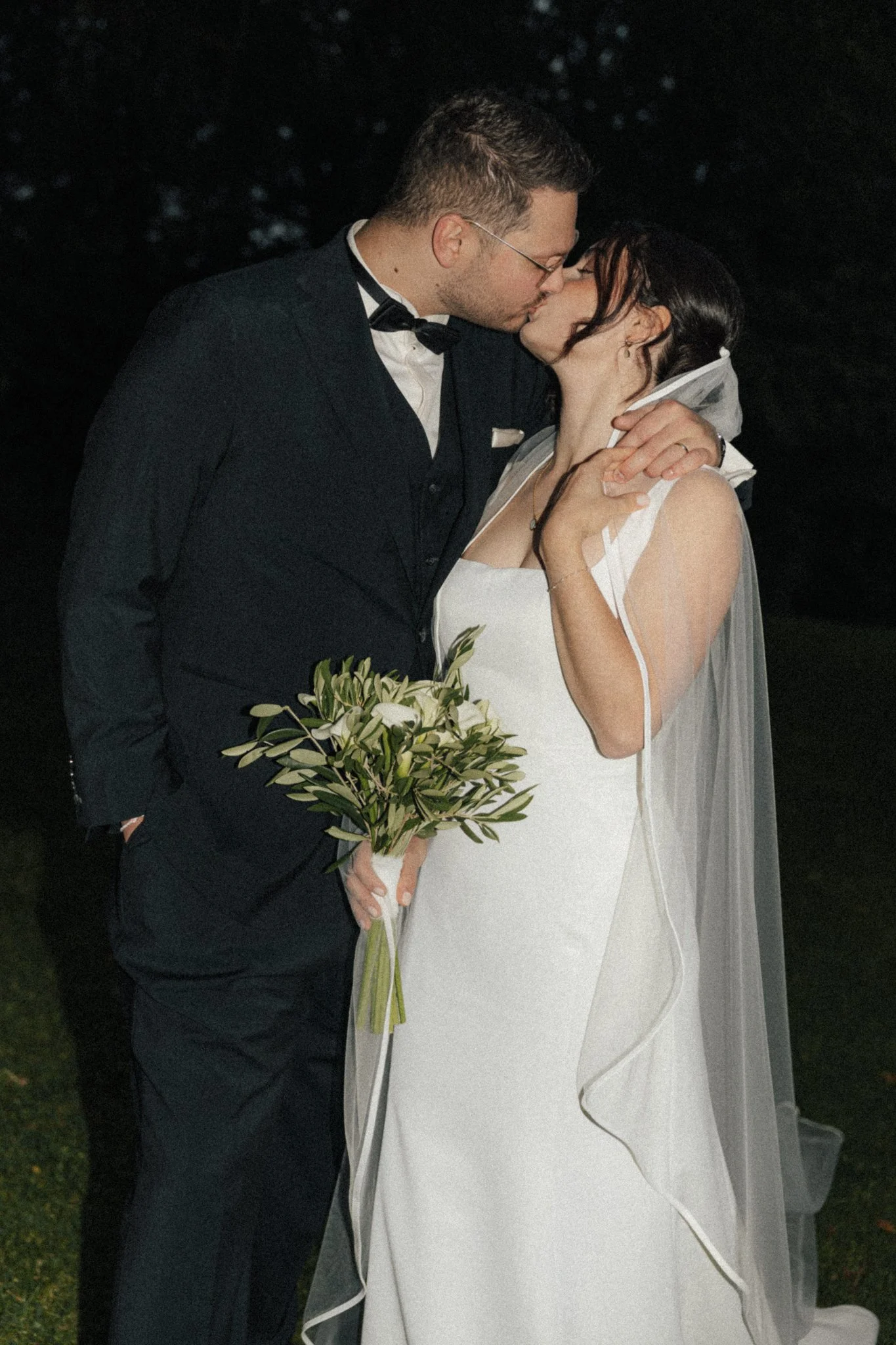 Ein Brautpaar bei der Hochzeitszeremonie, das sich küsst. Der Bräutigam trägt einen schwarzen Anzug mit Fliege, die Braut ein weißes Kleid mit Schleier, sie hält einen Blumenstrauß.