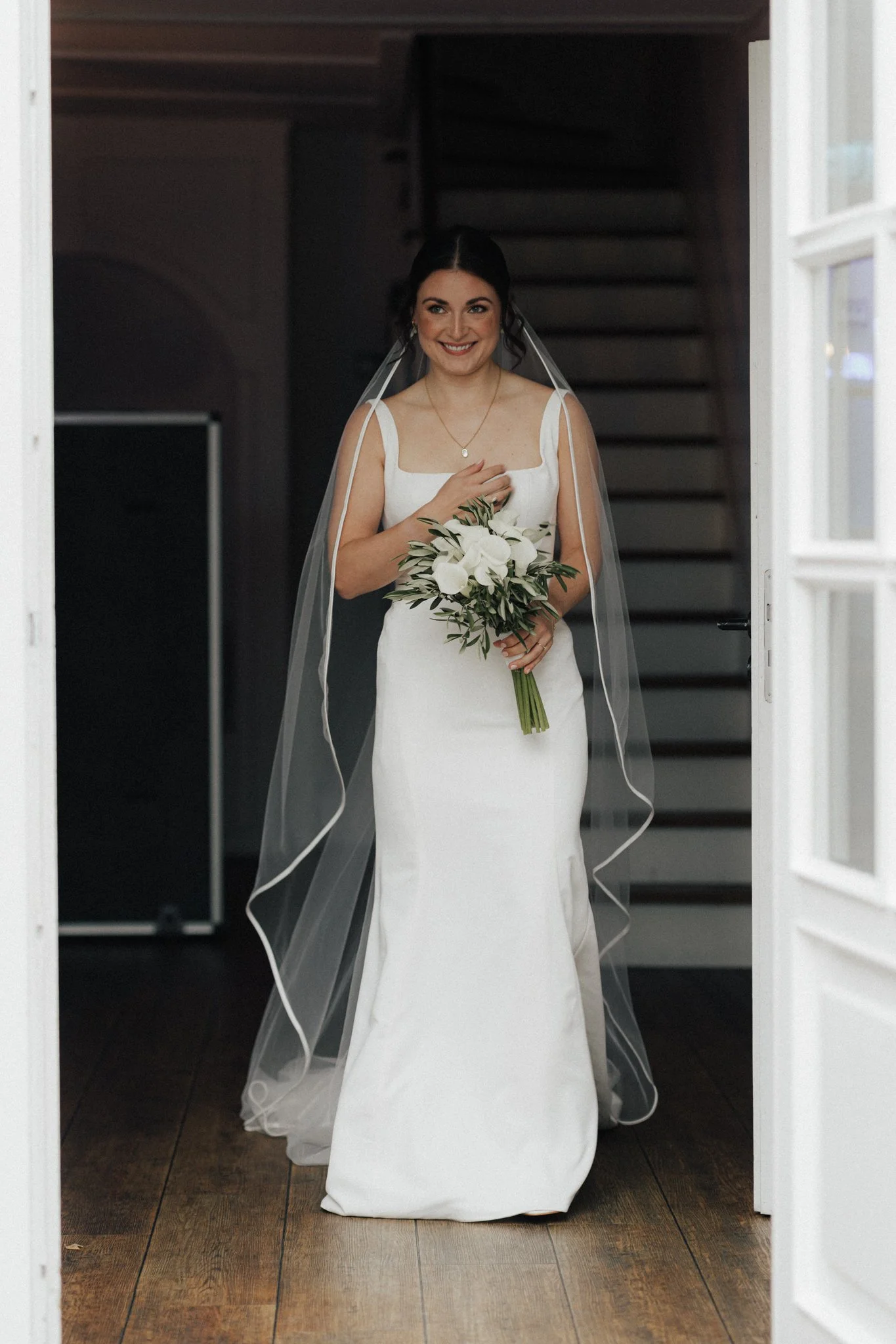 Braut mit Hochzeitskleid, Schleier, Hochzeitsstrauß, steht in einer Türöffnung im Innenbereich.