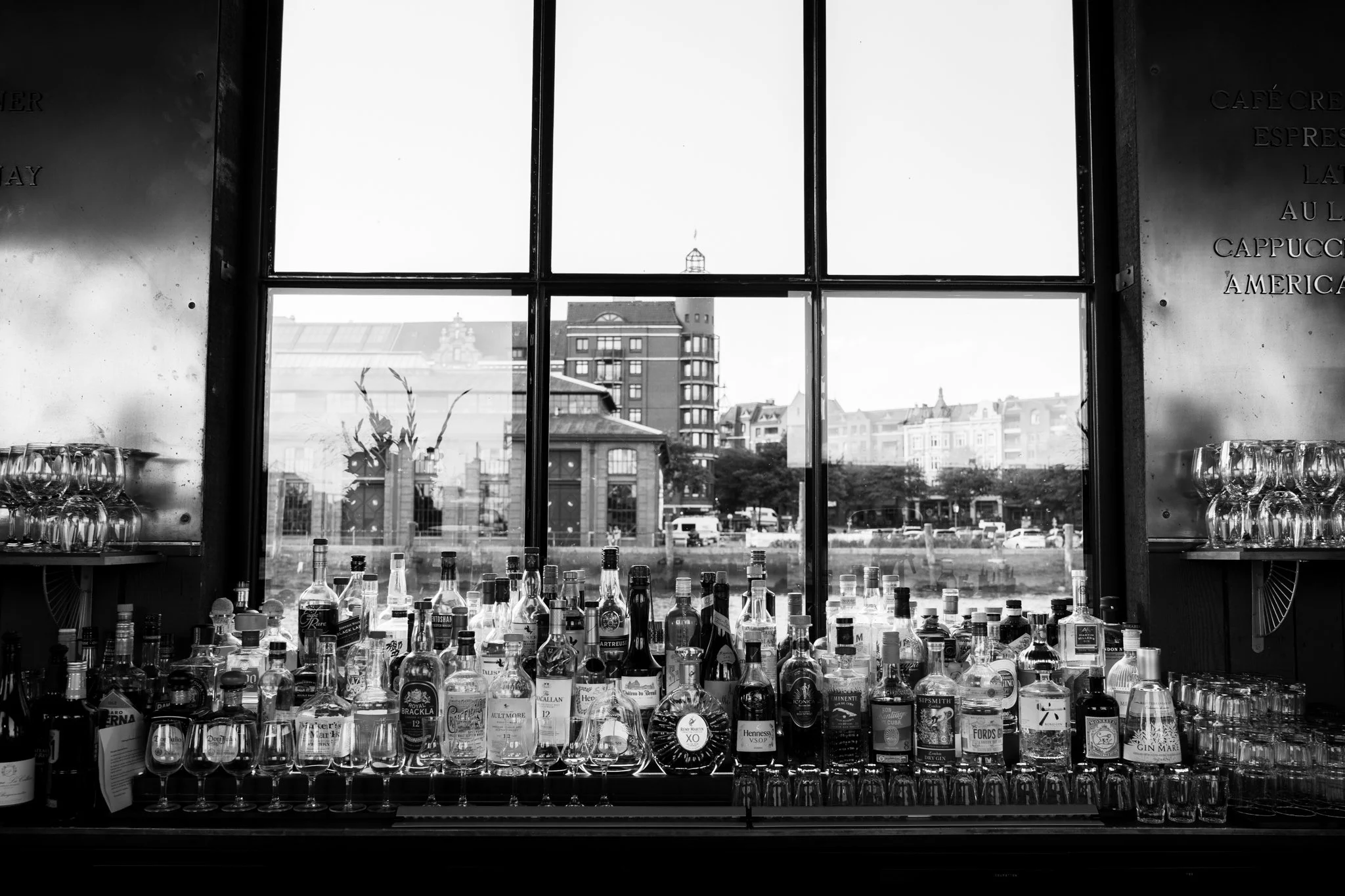 Blick durch ein großes Fenster auf eine Bar mit vielen Spirituosenflaschen und Gläsern im Vordergrund, städtische Gebäude im Hintergrund, Schwarz-Weiß-Fotografie.