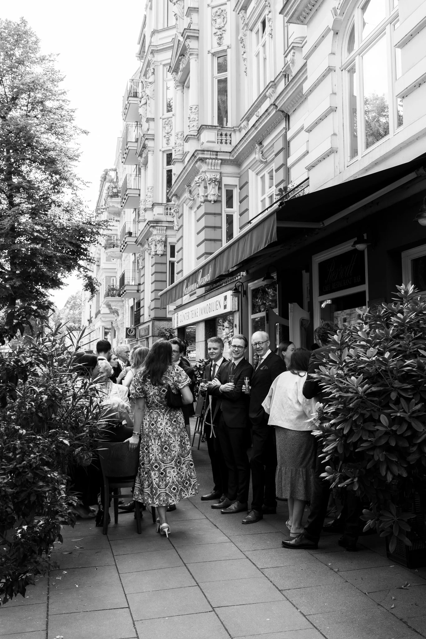 Menschen, die vor einem Café auf der Straße stehen, mit historischen Gebäuden im Hintergrund, in Schwarz-Weiß aufgenommen.