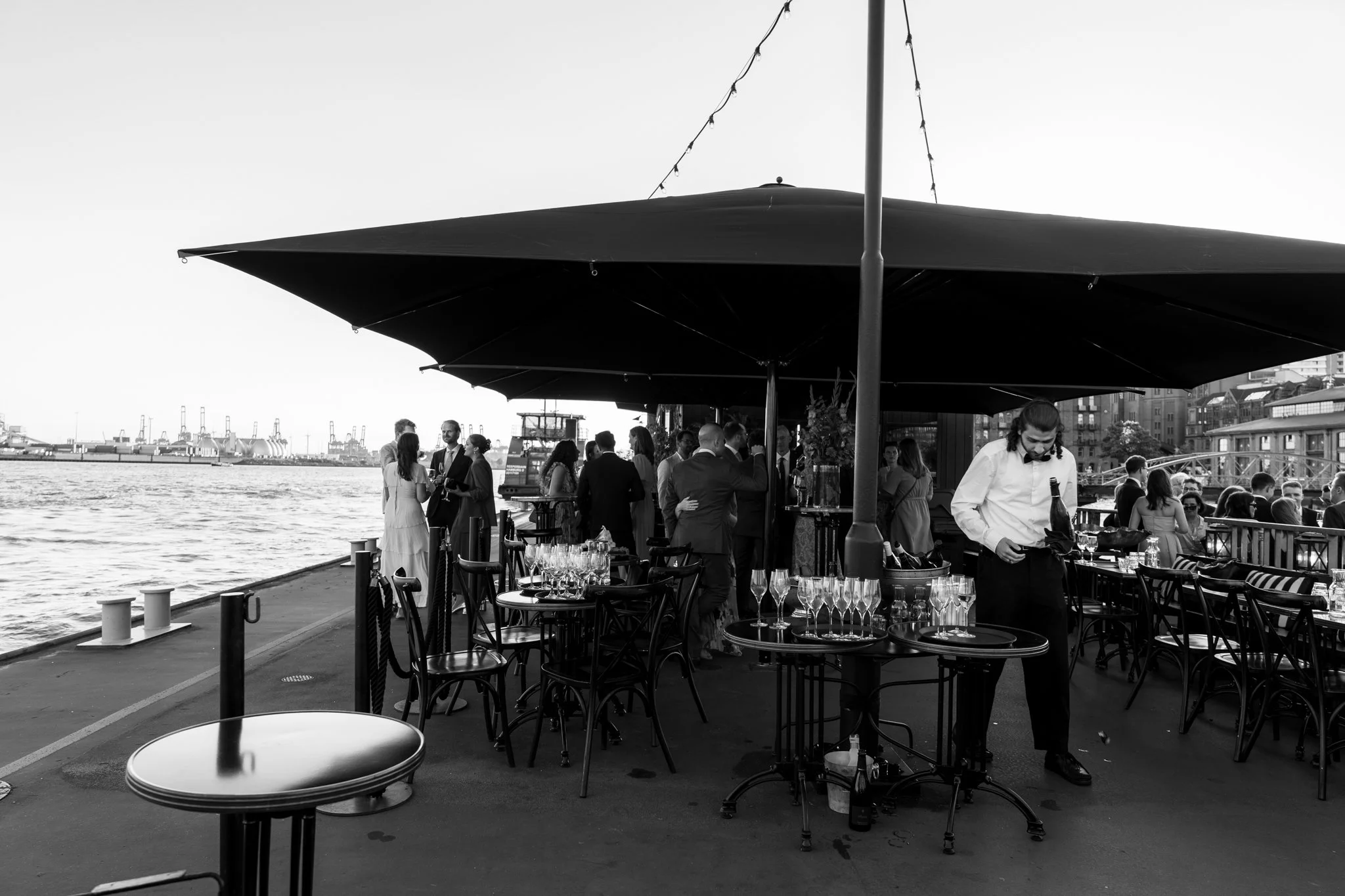 Blick auf eine elegante Abendveranstaltung an einem Fluss oder Hafen mit Tischen, Gläsern und Gästen, die sich unter einem großen Sonnenschirm versammeln, im Hintergrund eine Stadt.