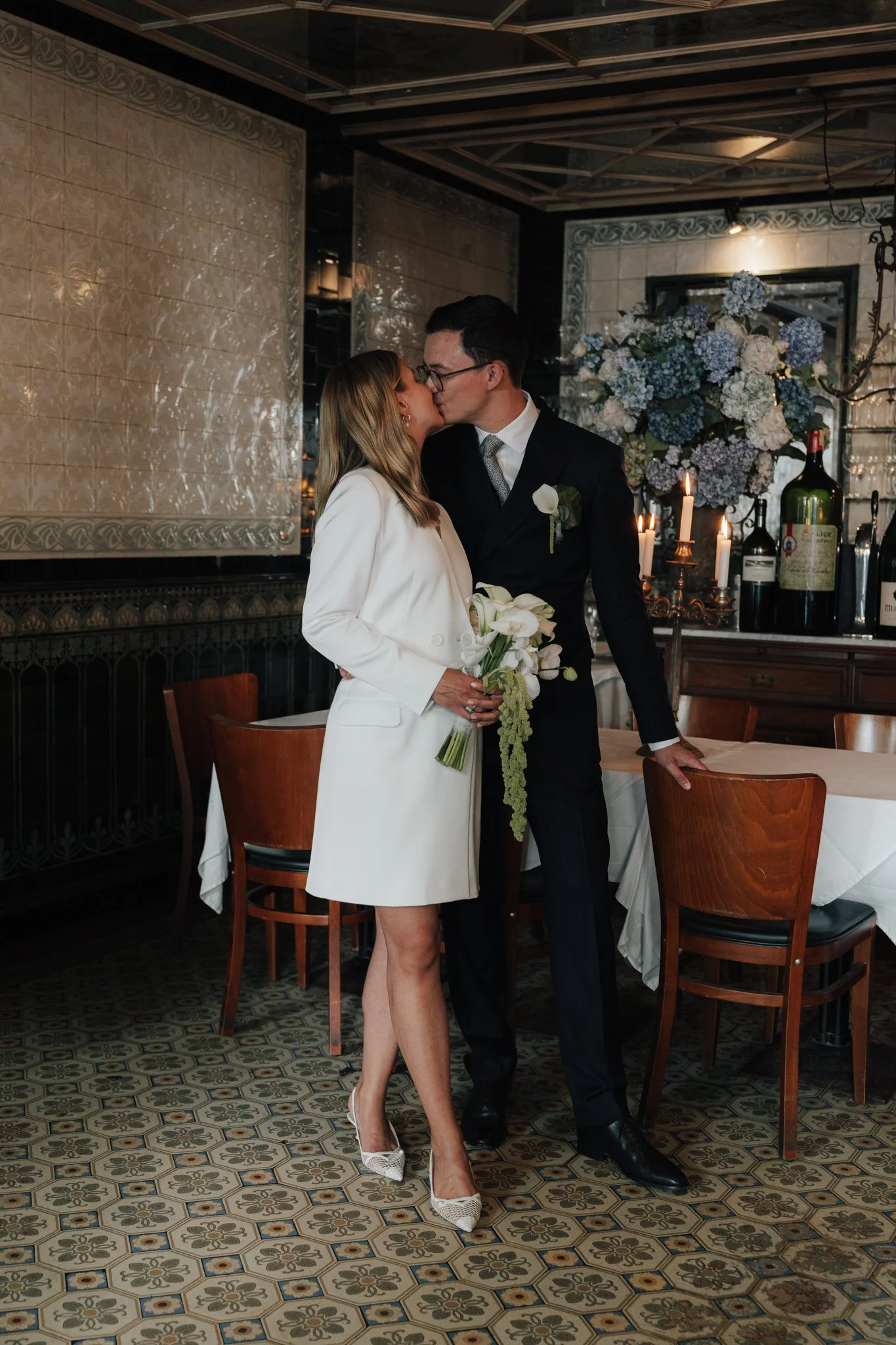 Ein Paar bei einer Hochzeit, sie küsst sich im Restaurant, sie trägt ein weißes Kleid, er trägt einen schwarzen Anzug.