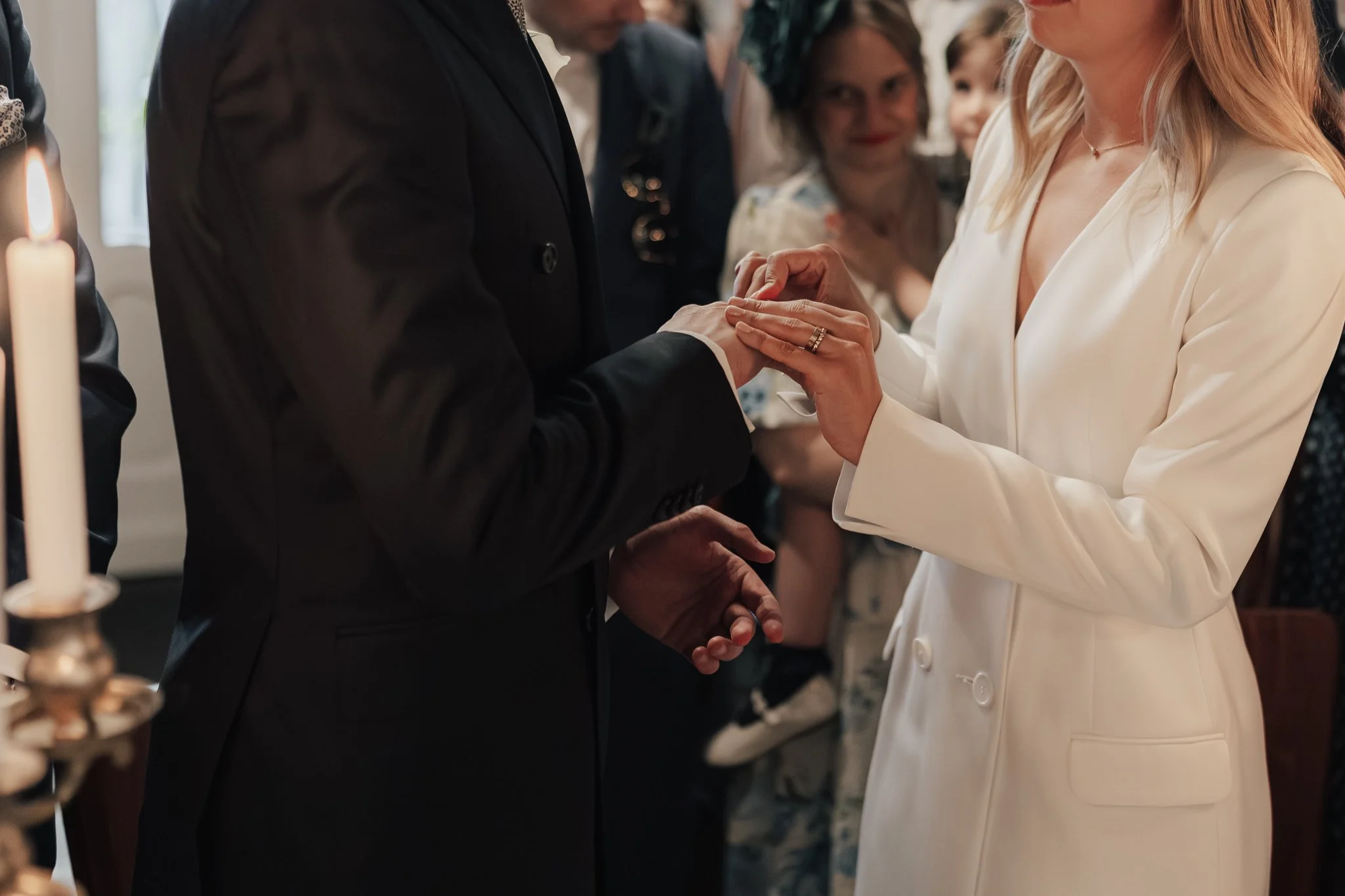 Eine Hochzeitszeremonie mit einem Brautpaar, das sich gegenseitig die Hände hält, während die Braut ein weißes Kleid trägt und der Bräutigam einen schwarzen Anzug trägt, umgeben von anwesenden Gästen.