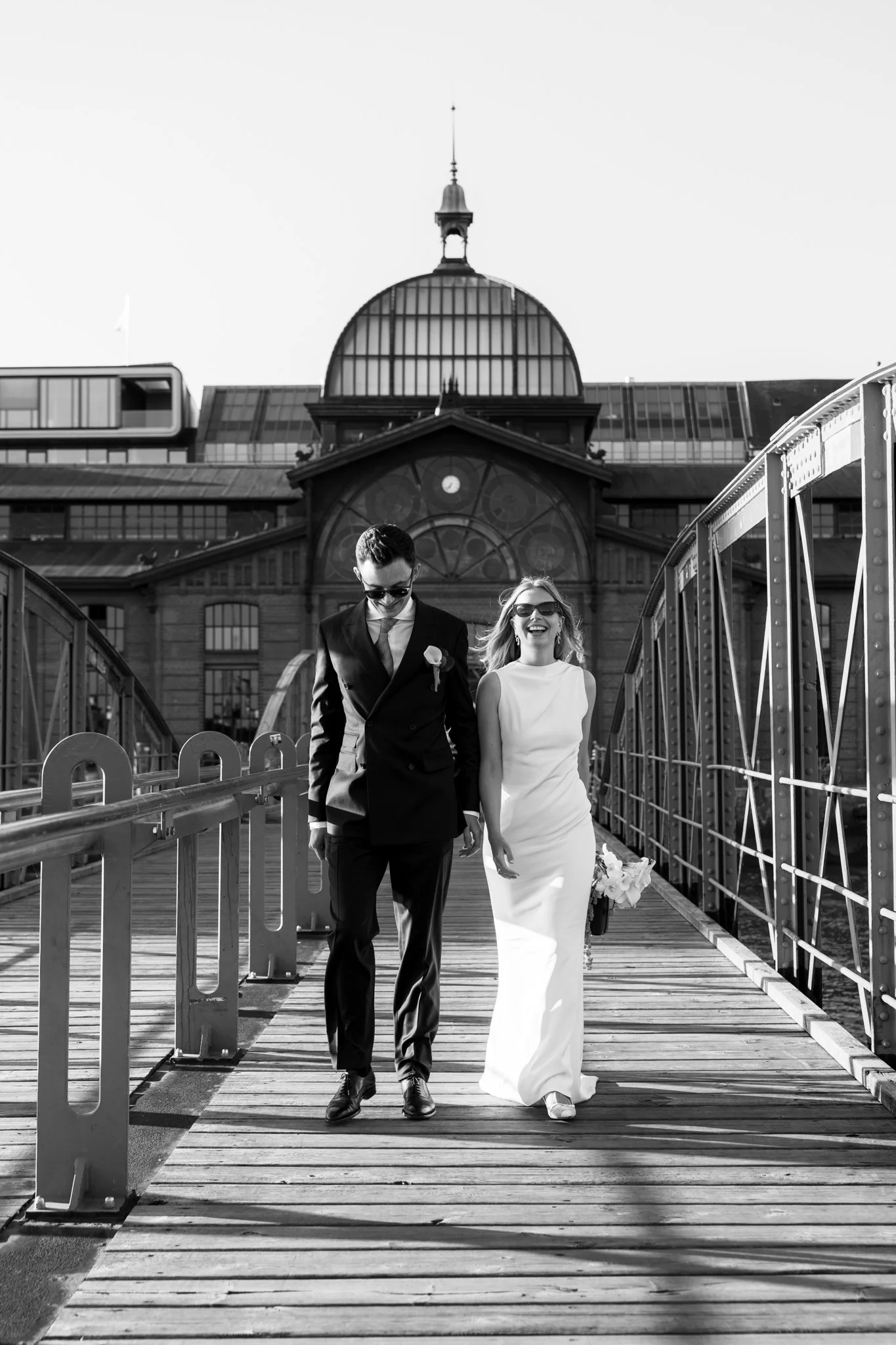 Ein glückliches Brautpaar in formeller Kleidung, unterwegs auf einer Brücke mit einer historischen Industriebau im Hintergrund, im Sonnenlicht, schwarz-weiß Fotografiefassung.