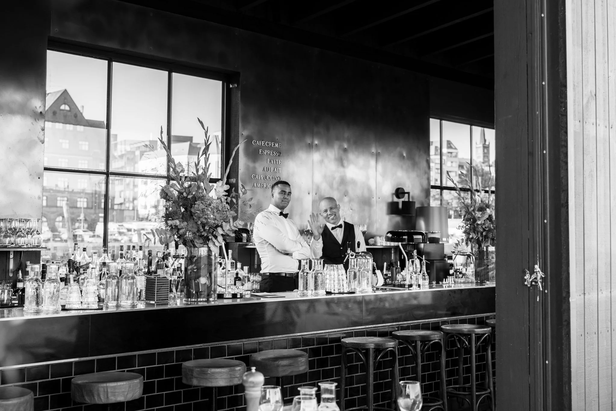 Zwei Barkeeper hinter einer Bar, umgeben von Gläsern, Flaschen und Blumen, in einem modernen Café oder Bar, mit großen Fenstern im Hintergrund.
