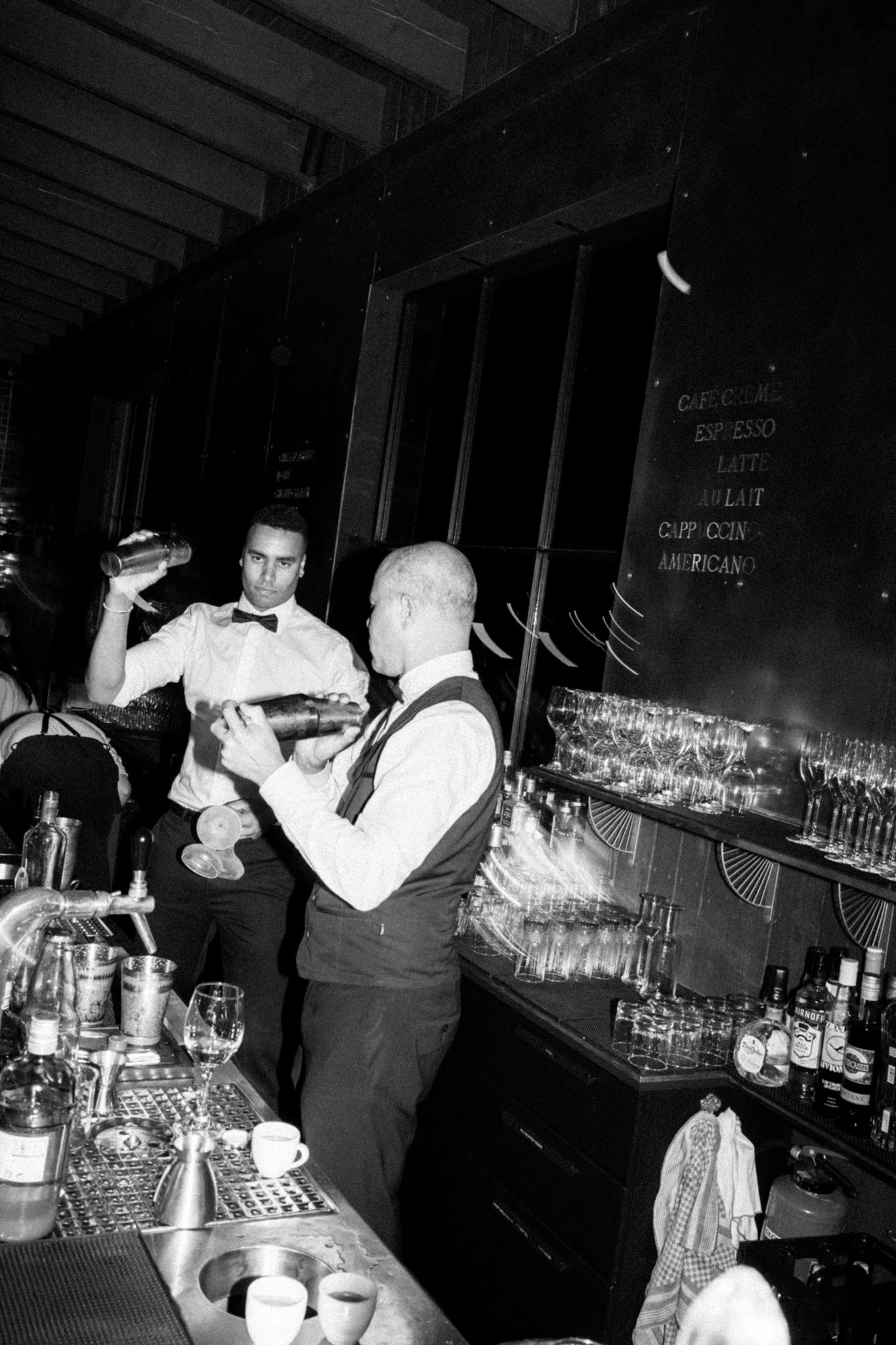 Zwei Baristas hinter einer Bar im Restaurant, einer mit einem Drink, der andere mit einer Flasche, in einem modernen, eleganten Raum.