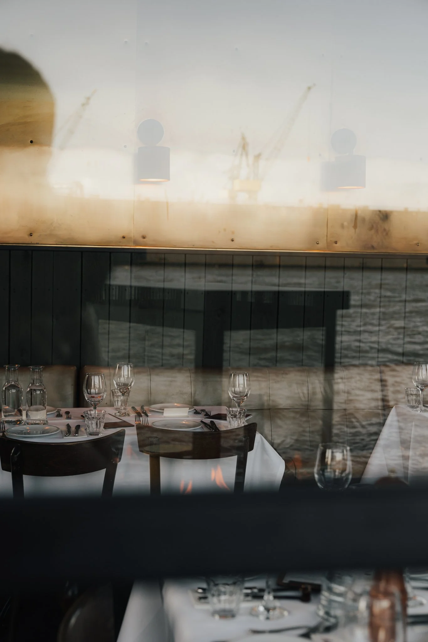 Ein Blick durch ein Fenster zeigt einen festlich gedeckten Restauranttisch mit weißen Tischtüchern, Wein- und Wassergläsern, Besteck, und Tellern. Im Hintergrund ist eine Wand mit einem Meeresblick und eine Wandbeleuchtung sichtbar.