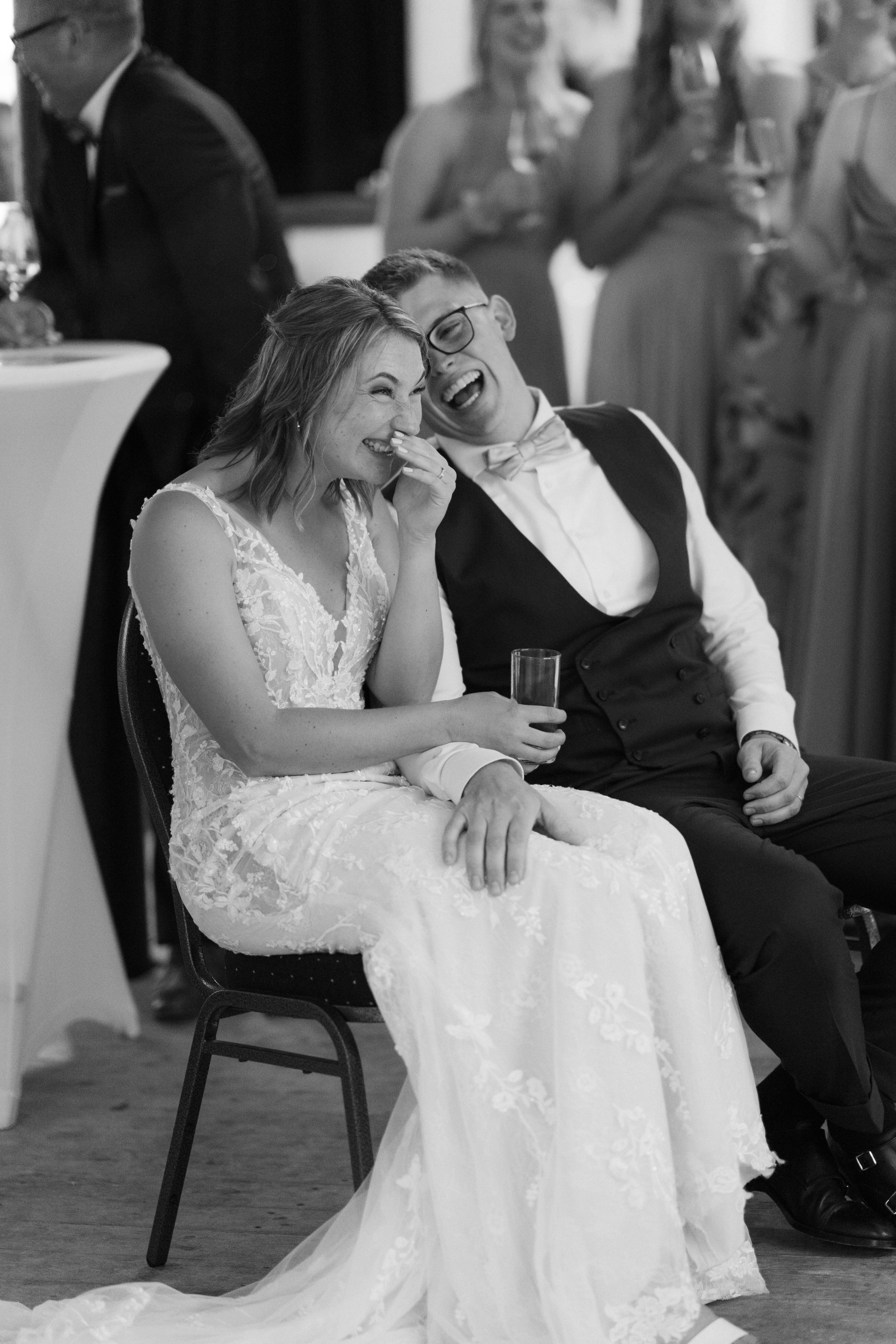 Braut und Bräutigam lachen bei einer Hochzeit, sitzend auf Stühlen, umgeben von Gästen.