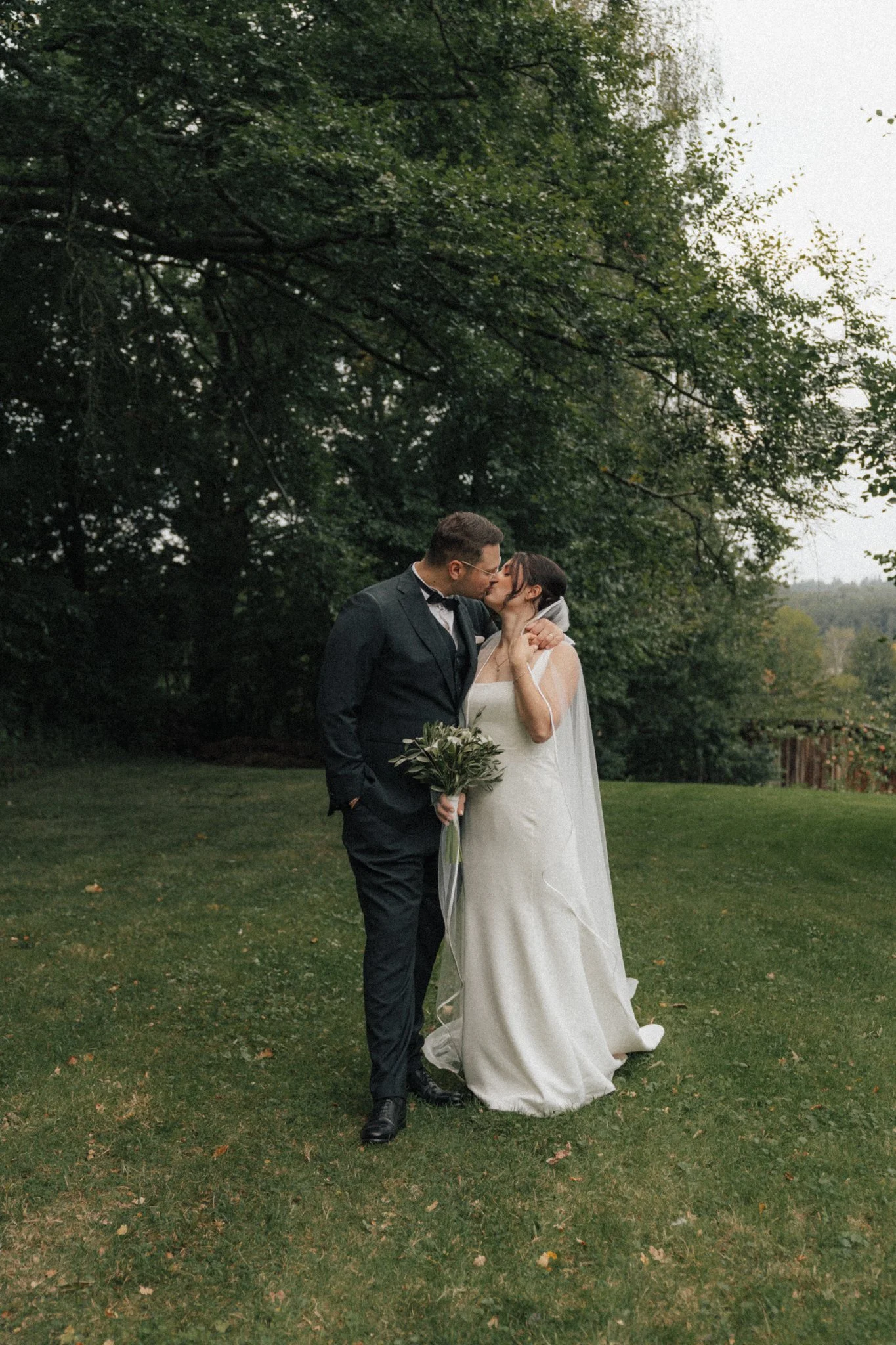Ein frisch verheiratetes Paar, das sich küsst, steht im Grünen unter einem Baum im Freien.