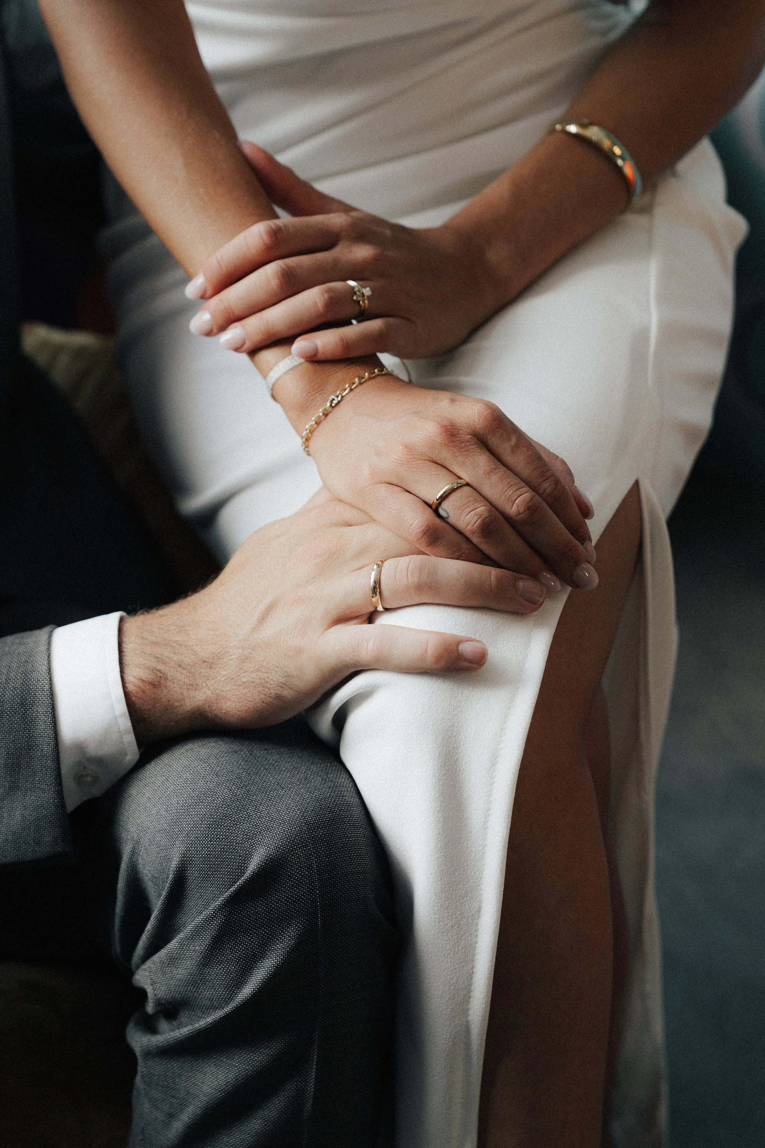 Nahaufnahme von vier Händen, die auf den Knien einer Person ruhen, mit mehreren Ringen und Schmuck, in einem formellen oder besonderen Anlass, wahrscheinlich Hochzeit oder Feierlichkeiten.