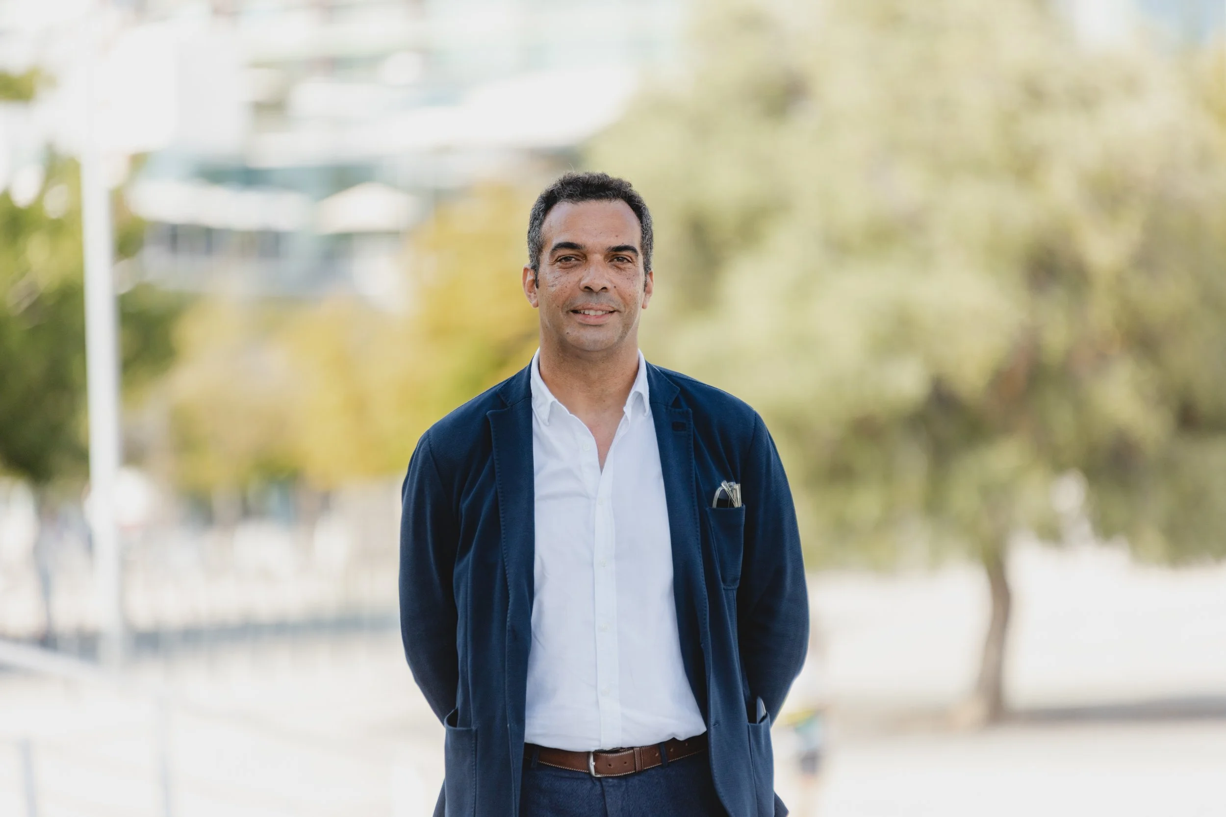 A man in a navy blazer and white shirt standing outdoors with trees and buildings in the background.