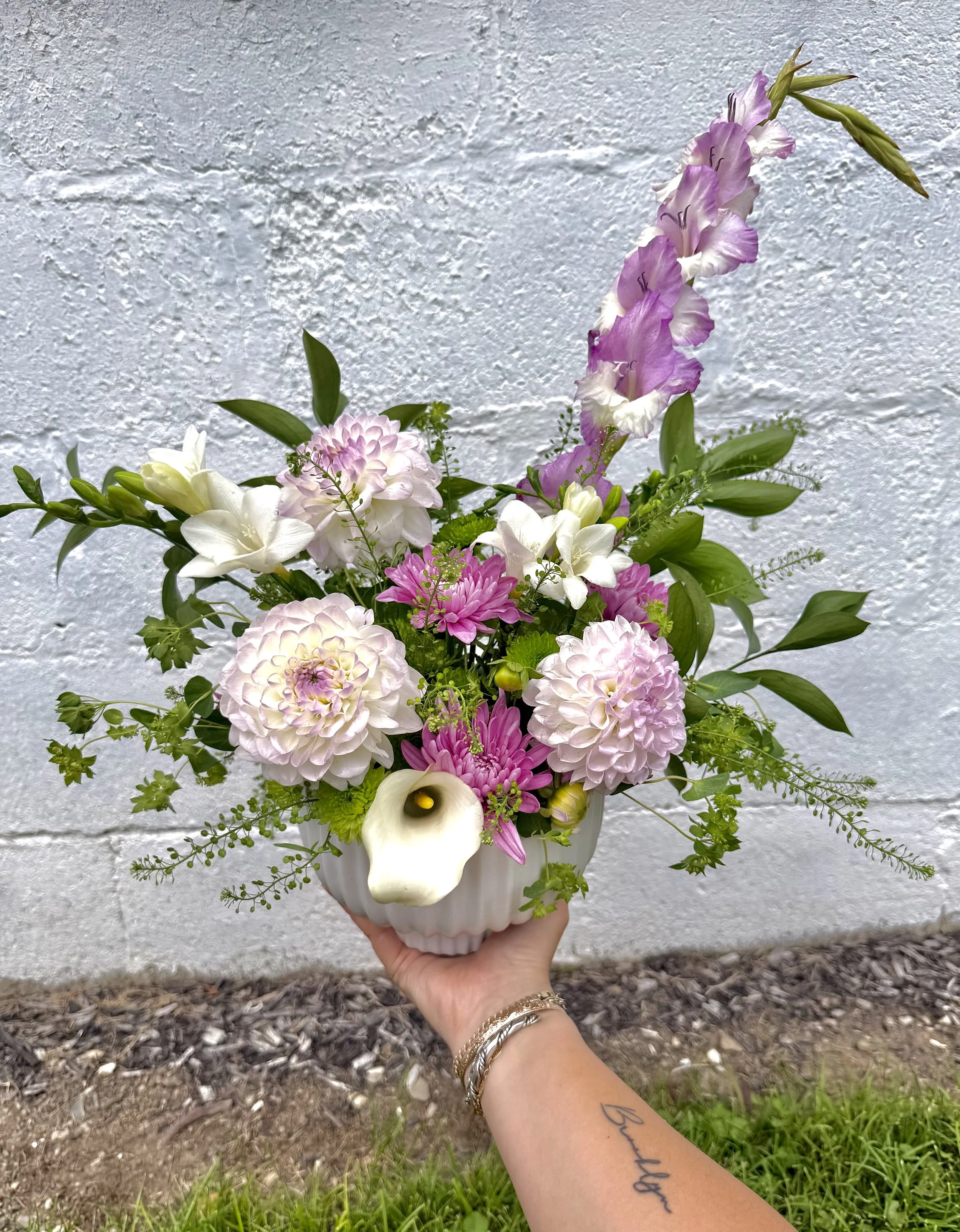 A hand holding a white vase with an arrangement of pink, white, and purple flowers, including calla lilies, roses, and other mixed blooms, against a white textured brick wall background.