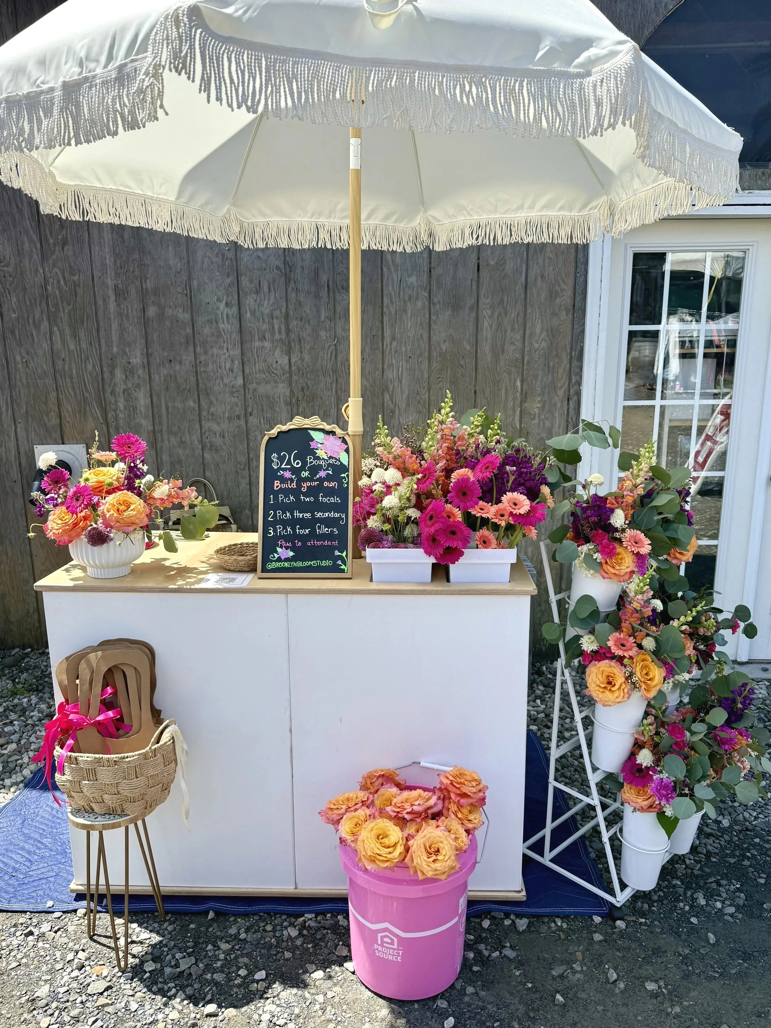 Flower bar with pink, purple, and peach blooms, a chalkboard sign, and a white table under a large white parasol. A wicker basket with a pink ribbon, and a pink bucket filled with peach roses are also visible.