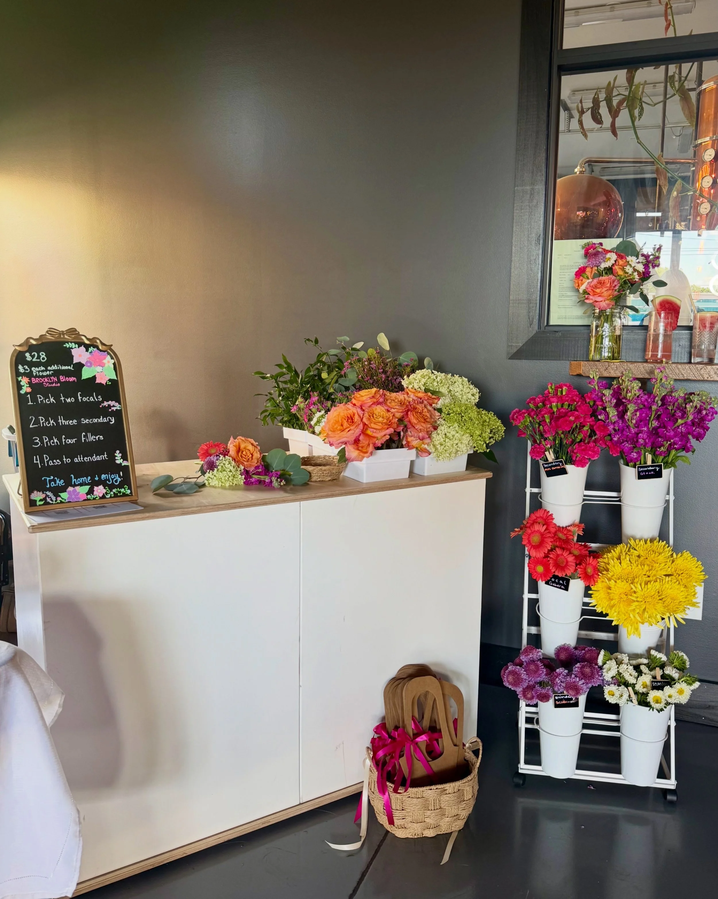 Display of various colorful flowers in vases and containers, with a blackboard sign listing flower prices and details, situated indoors near a window and a white shelving unit holding potted flowers.