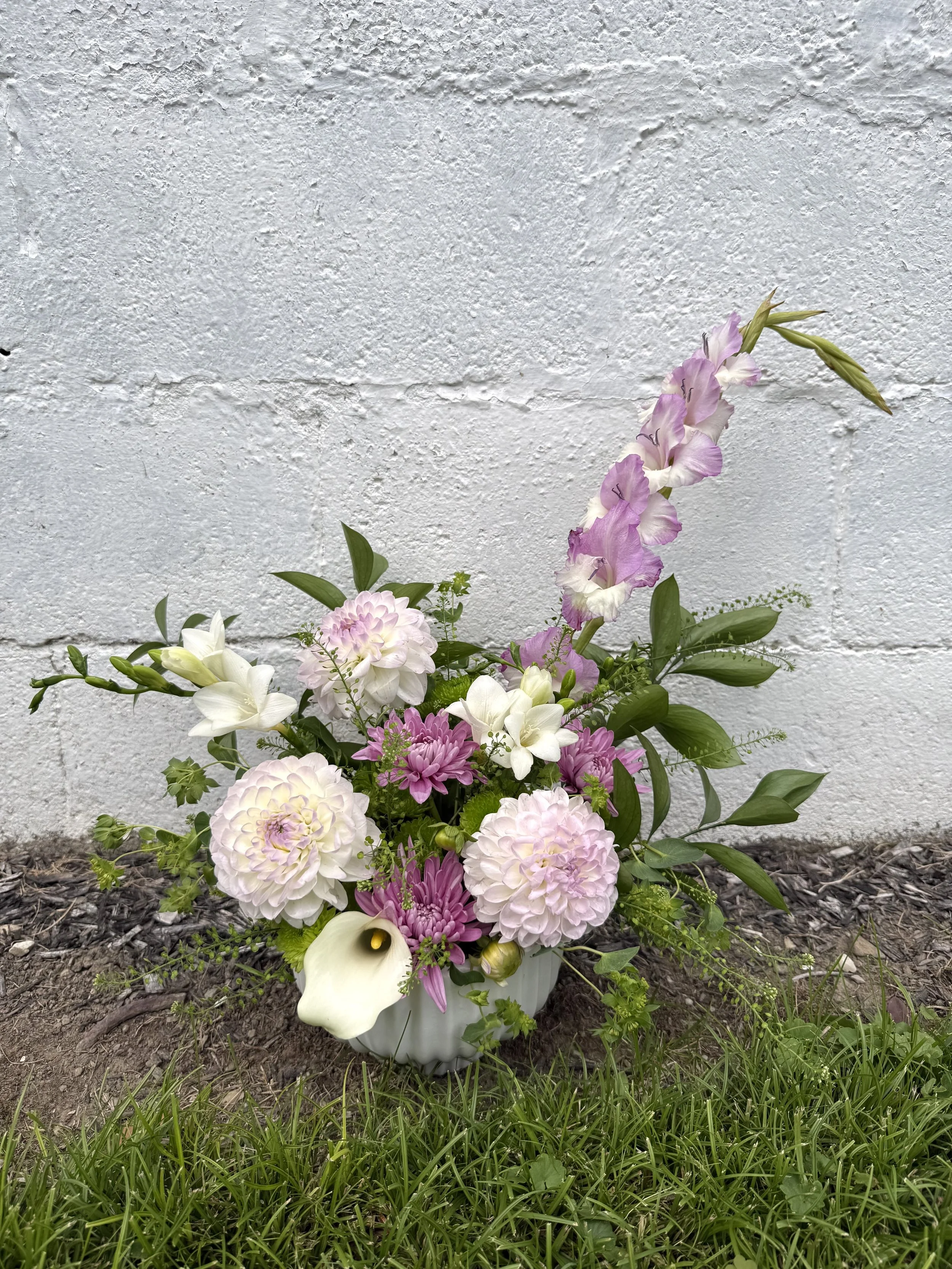 A flower arrangement in a white vase against a white brick wall, with various pink, white, and purple flowers and green foliage, placed outdoors on soil and grass.