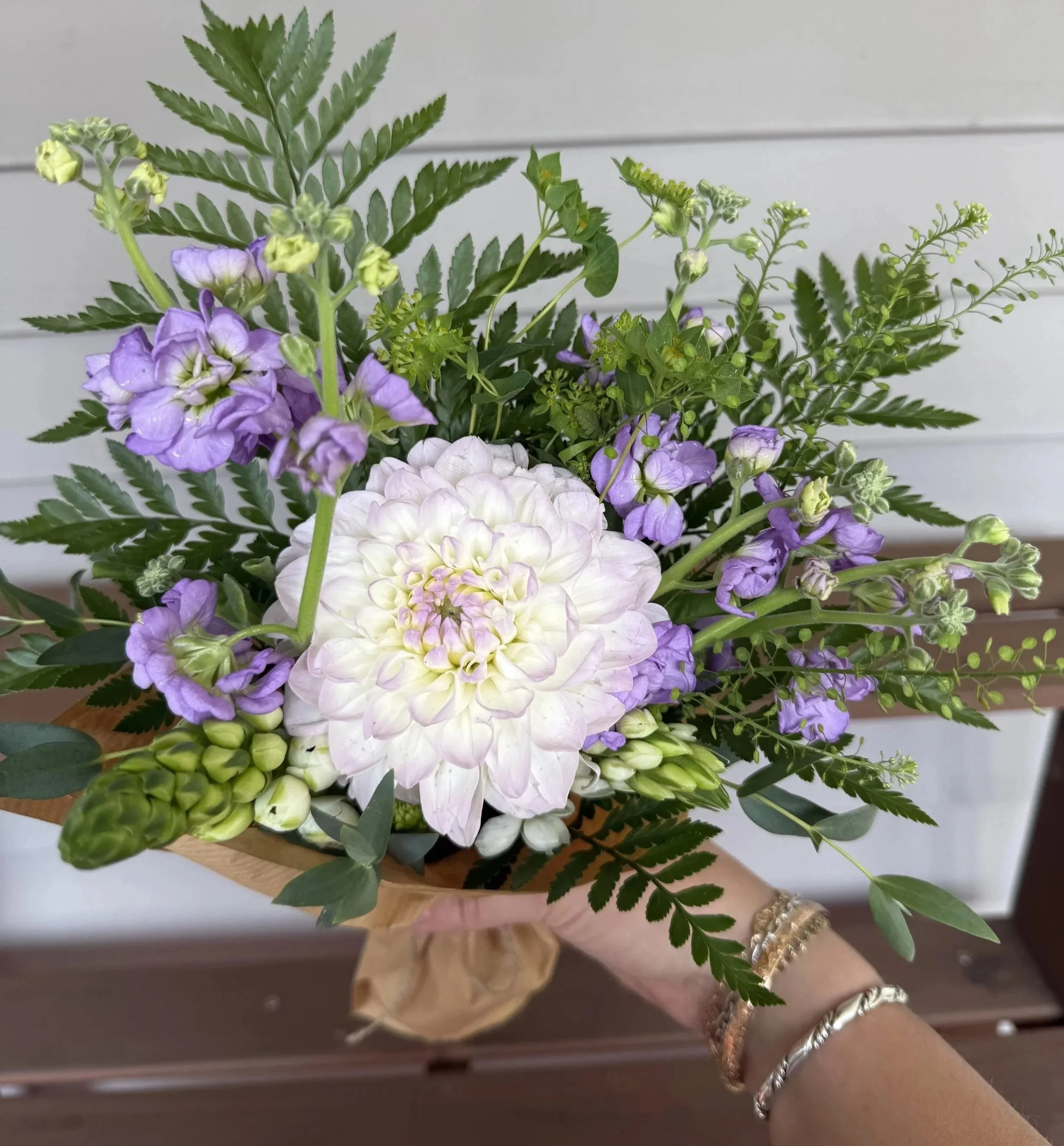 A person's arm with 3 silver bracelets holding a bouquet of purple, white, and green flowering plants with green fern-like leaves, on a white horizontal-paneled wall background.