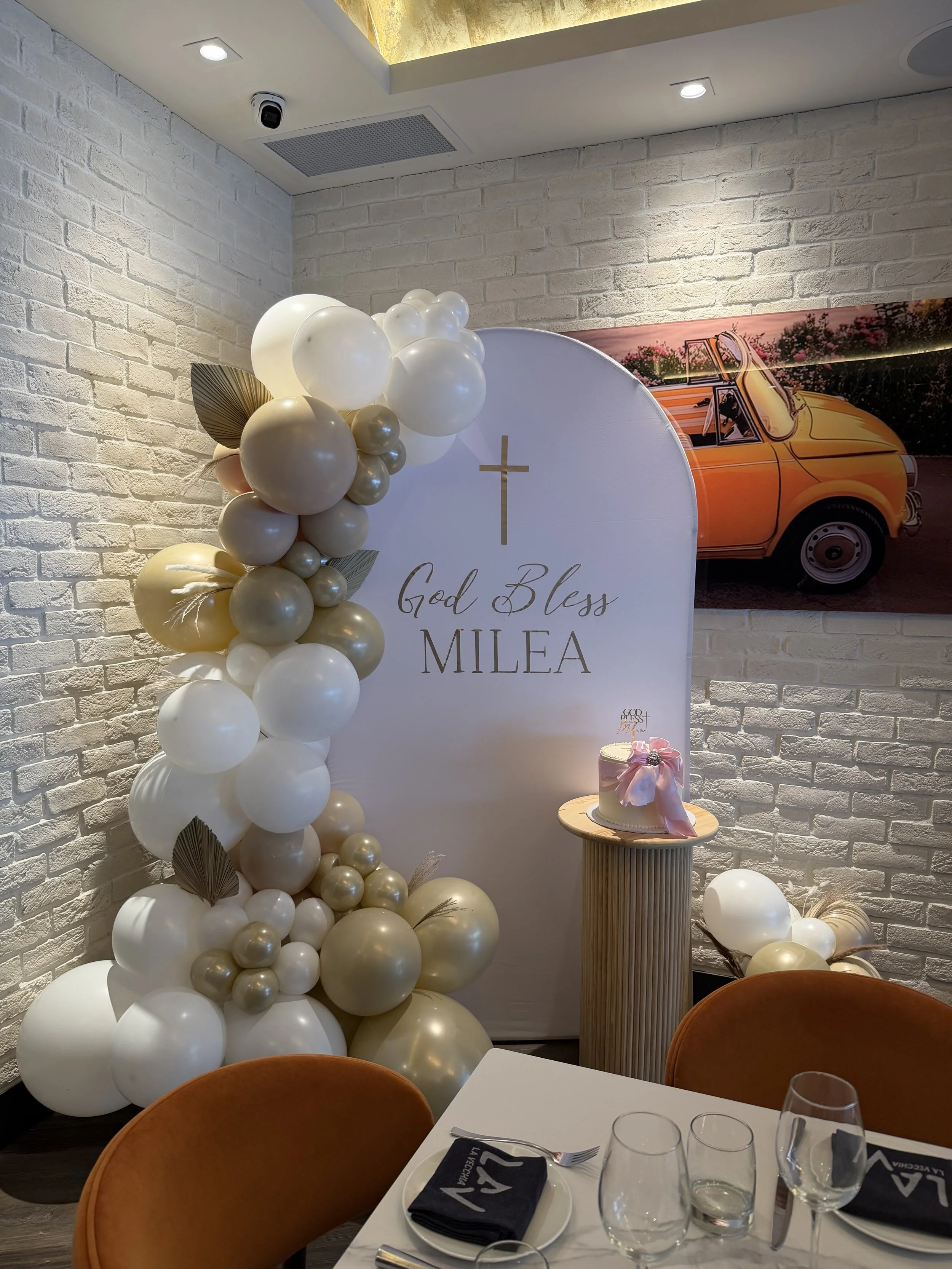 Decorative display for a celebration with balloons, a white backdrop with a cross and the words 'God Bless Millea,' a small cake with a pink ribbon, and a background image of a vintage yellow car on a wall with white brick texture.