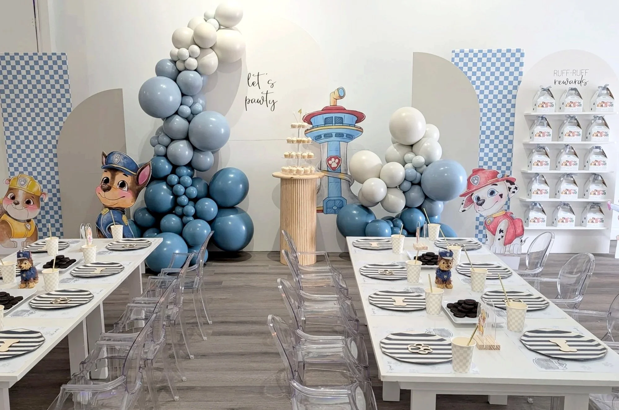 Party room decorated with blue and white balloons, Paw Patrol-themed character cutouts, and a table set for a celebration with striped plates, cups, and themed party favors.