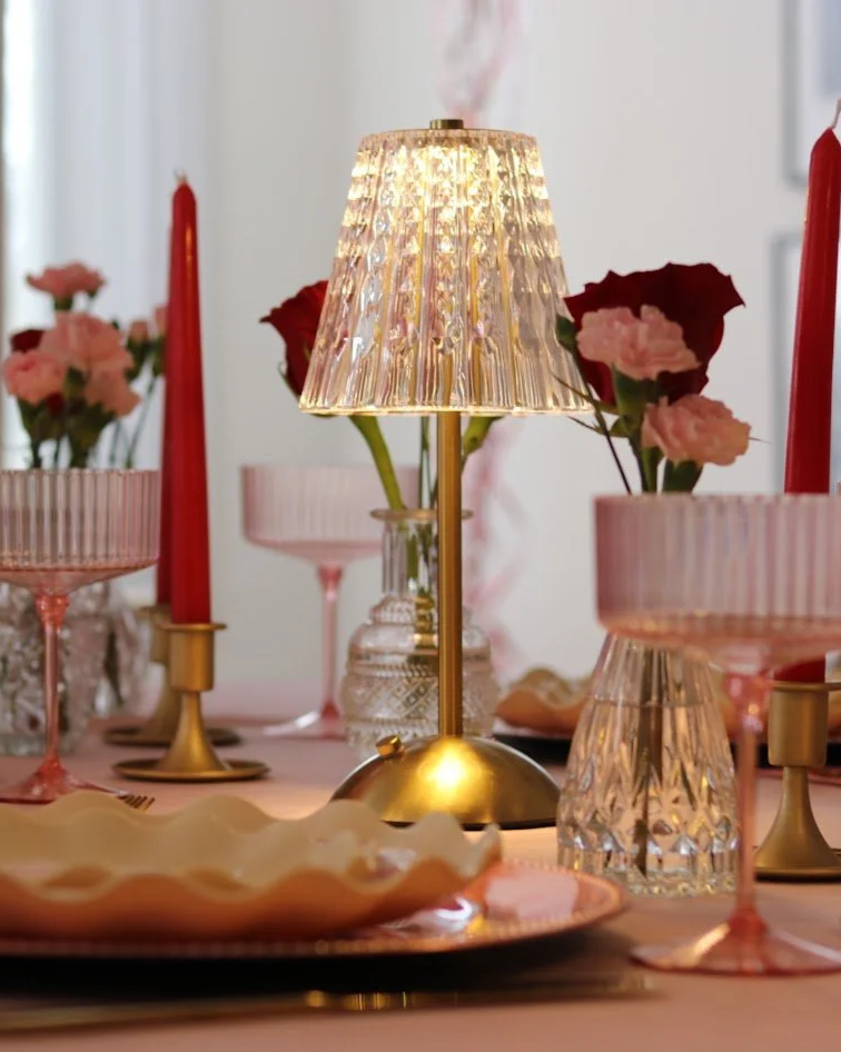 A decorated dining table with a pink tablecloth, featuring a gold and glass table lamp, pink glass wine glasses, pink candlesticks, pink flowers in vases, a decorative shell plate, and various glassware and cutlery.