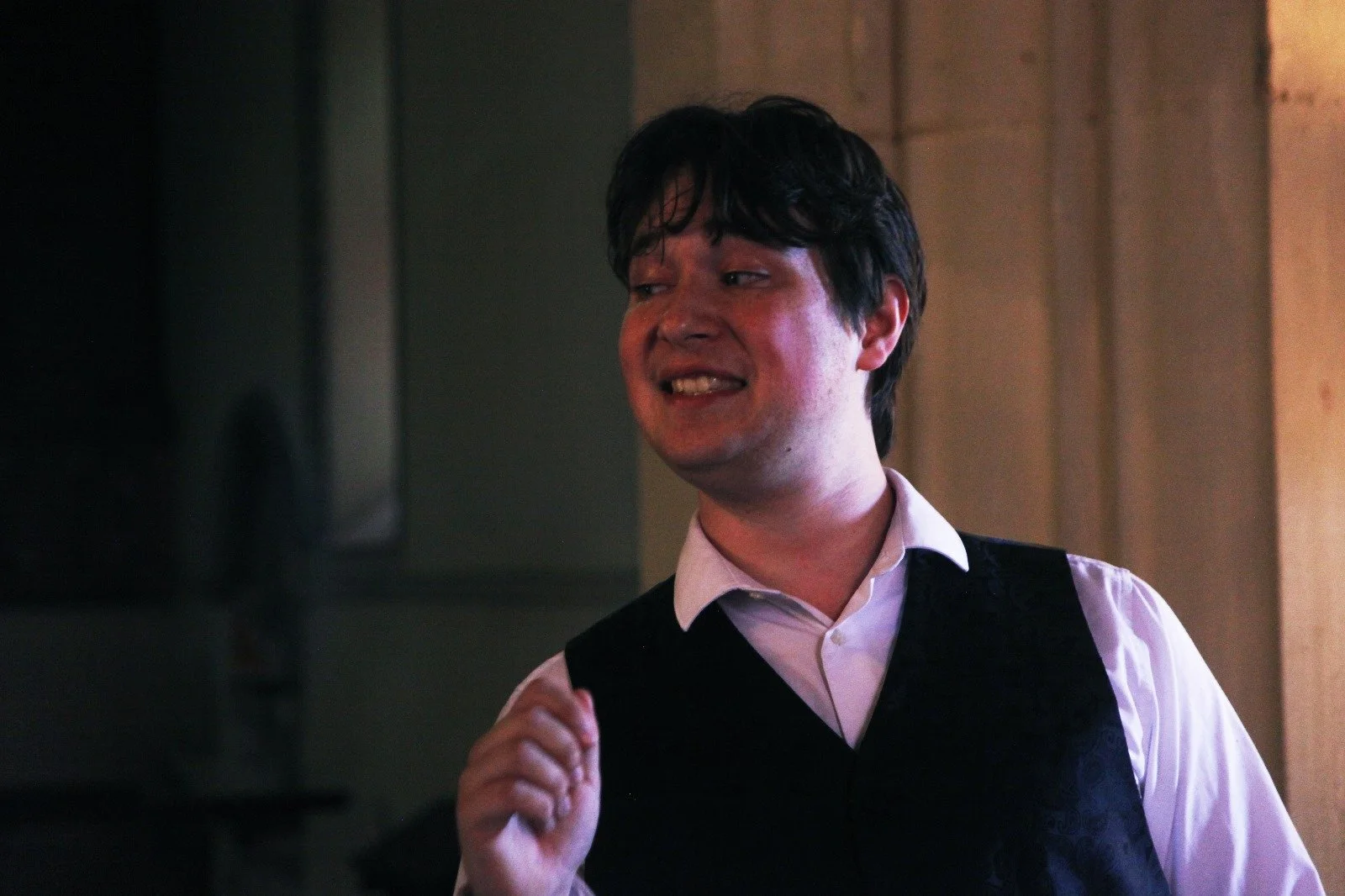A young man with dark, slightly messy hair, wearing a white shirt and a black vest, making a facial expression with bared teeth and slightly squinted eyes, in an indoor setting with warm lighting.