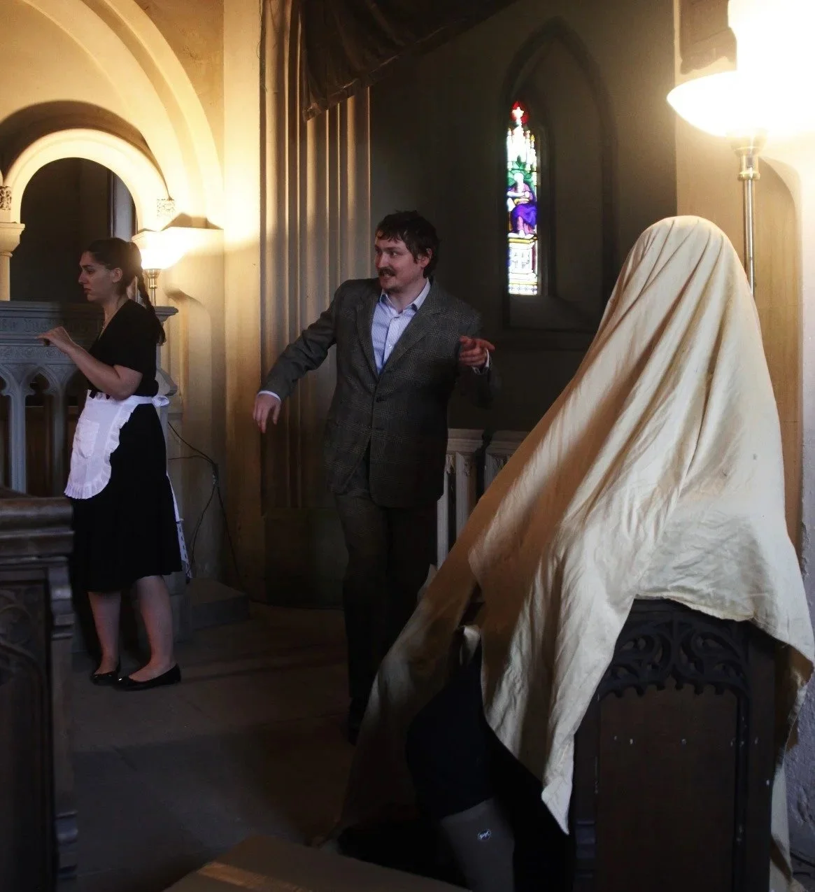 People inside a church. A woman dressed as a maid, another person covered with a cloth, and a man in a suit are present. The background shows stained glass windows.