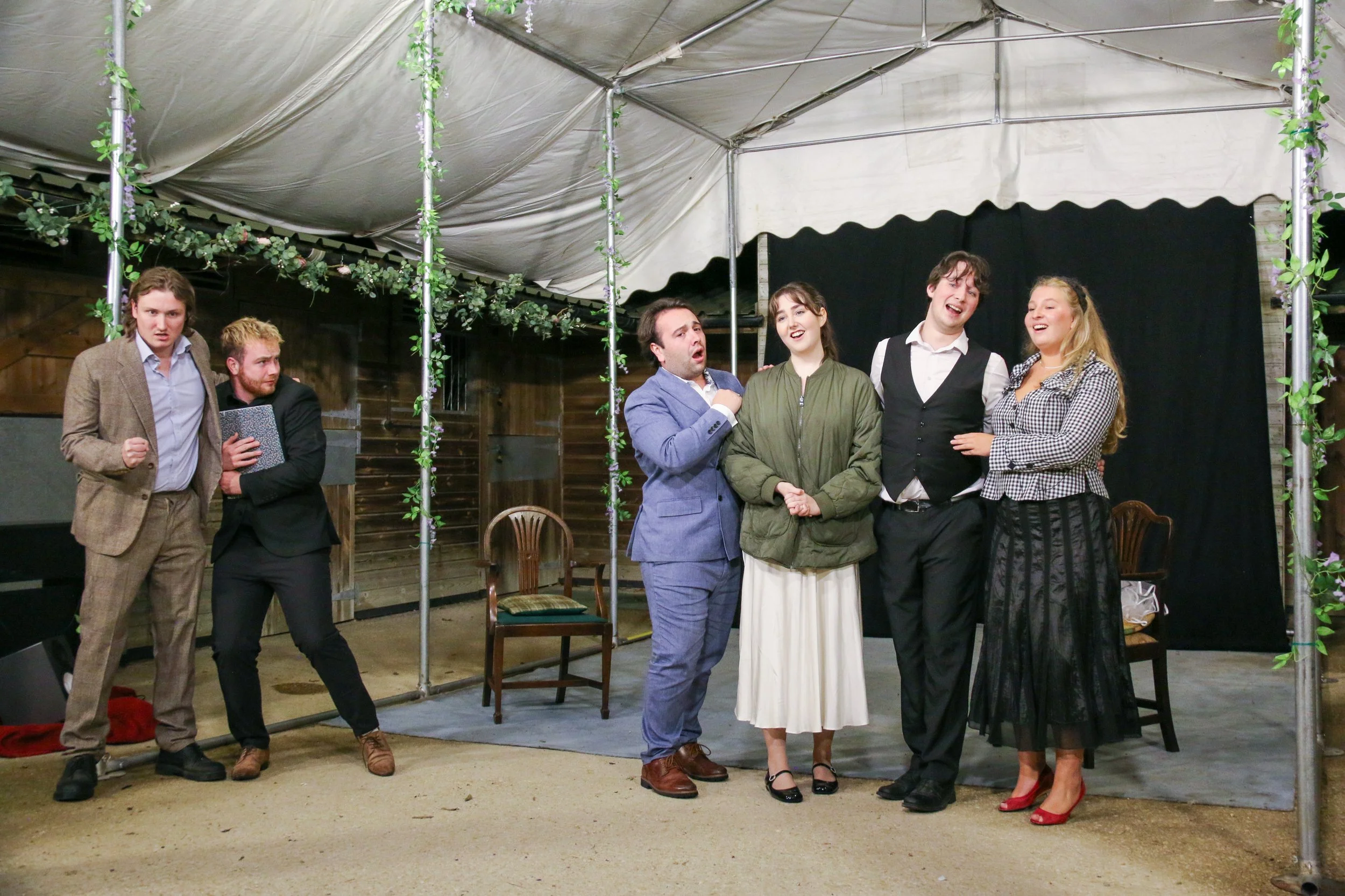 Group of six actors performing on a stage with a black backdrop and decorated with greenery, under a tent with artificial grass on the floor.