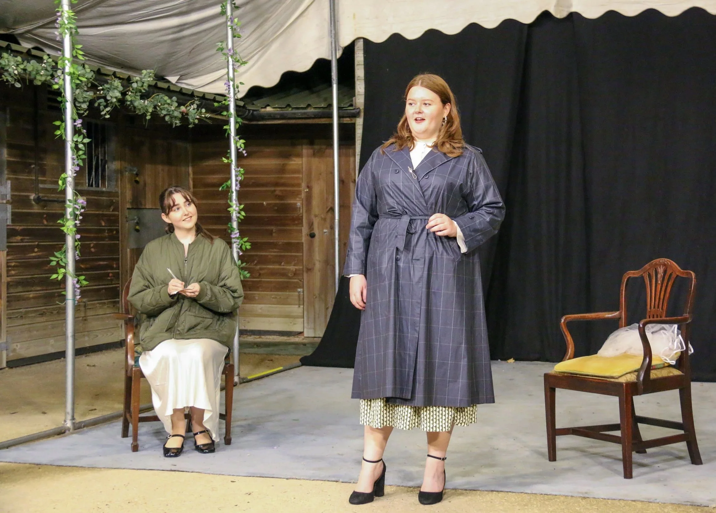 Two women performing on a small stage. One woman stands in the foreground wearing a checked dress with a dark blue coat, and the other woman sits in the background wearing a green jacket and white dress, taking notes. The stage has wooden walls, and there are two chairs, one empty and one with a bag on it, and a black curtain backdrop.
