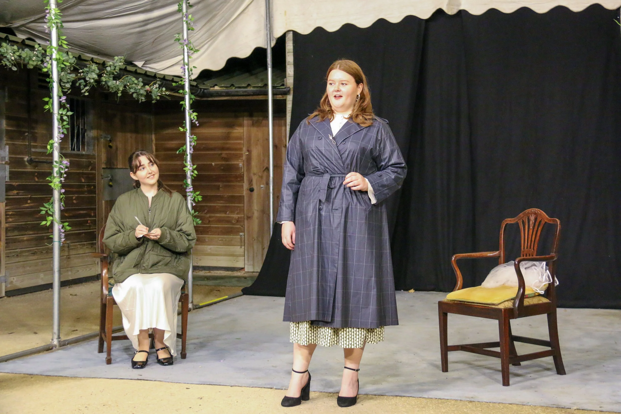 Two women on stage, one standing and one sitting, in what appears to be a theatrical performance. The standing woman has red hair and is wearing a dark plaid trench coat with high heels. The seated woman has dark hair, wearing a green jacket and a wh