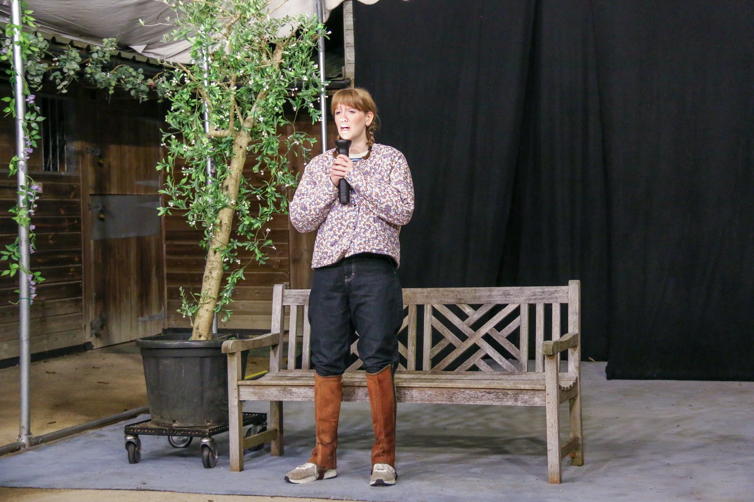 A woman holding a microphone standing on a stage with a wooden bench, a potted plant, and a black curtain backdrop.