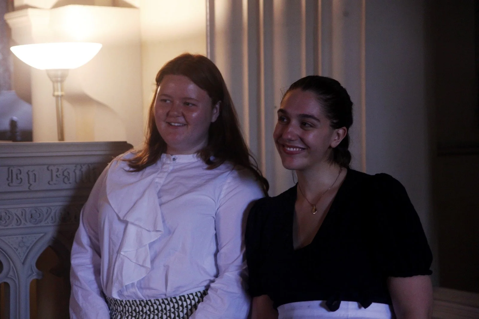 Two women standing indoors in front of a lit lamp and a decorative fireplace, smiling and posing for a photo.
