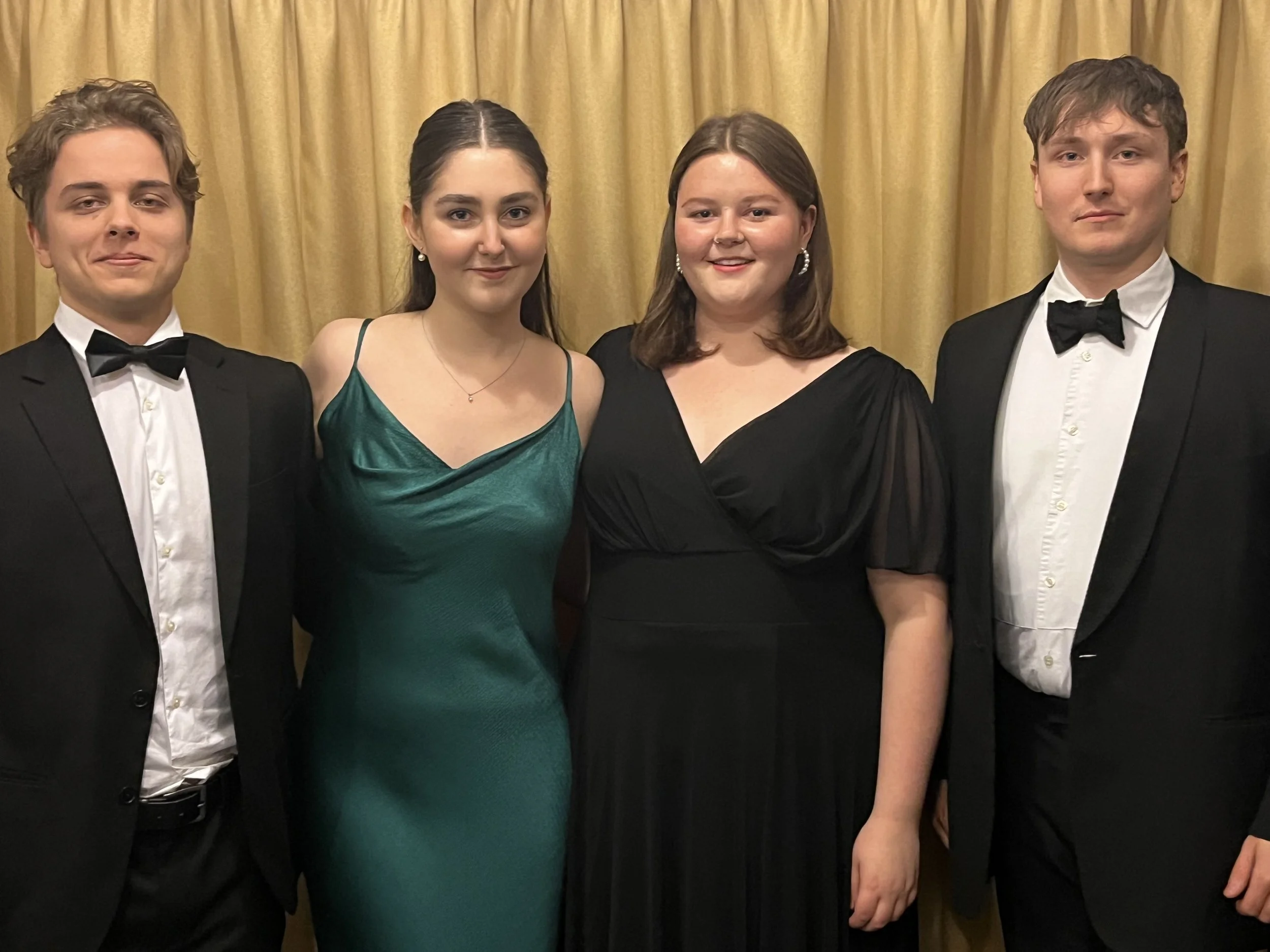 Four young adults dressed in formal attire, standing in front of a yellow curtain, attending a formal event or celebration.