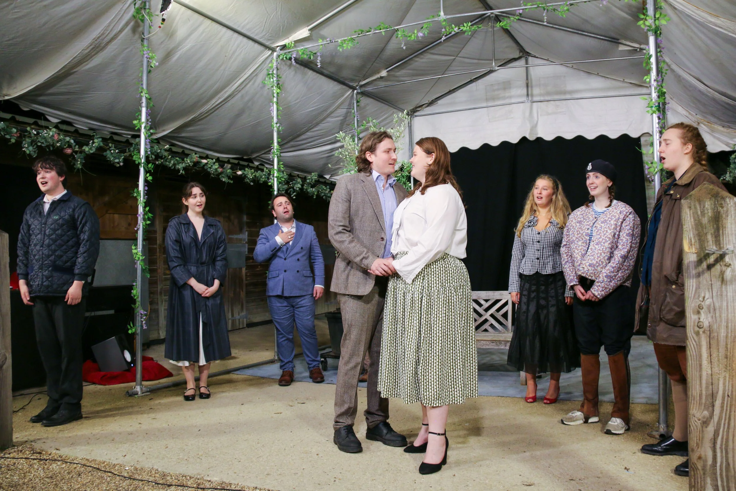 A group of actors performing a scene on a stage with a tent overhead, surrounded by decorative greenery.