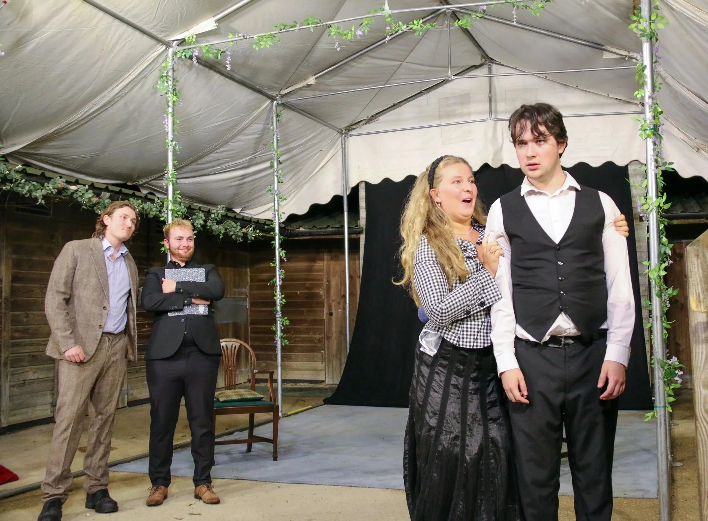 Four people performing a theatrical play under a tent; a woman appears emotional while holding a young man's arm, and two men are watching nearby, one smiling with arms crossed and the other observing thoughtfully.
