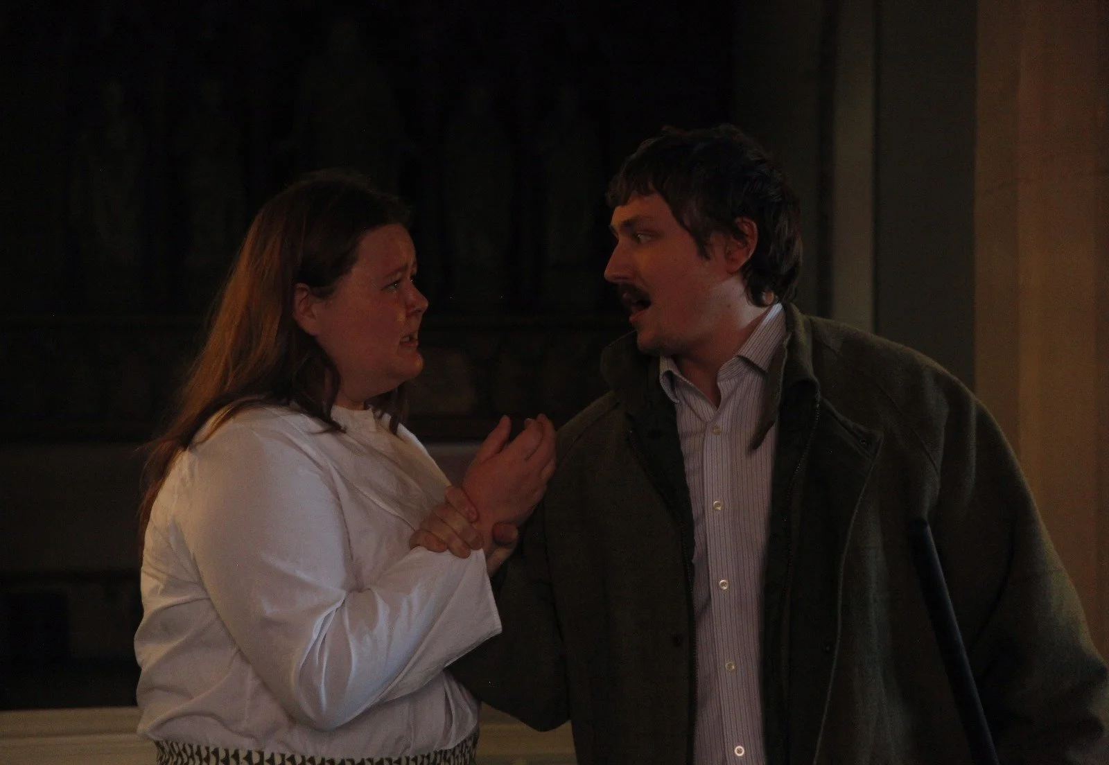 A woman in a white shirt appears emotional, holding her hands together, while a man in a dark jacket and striped shirt talks to her with concern. They are indoors, in a dimly lit room.