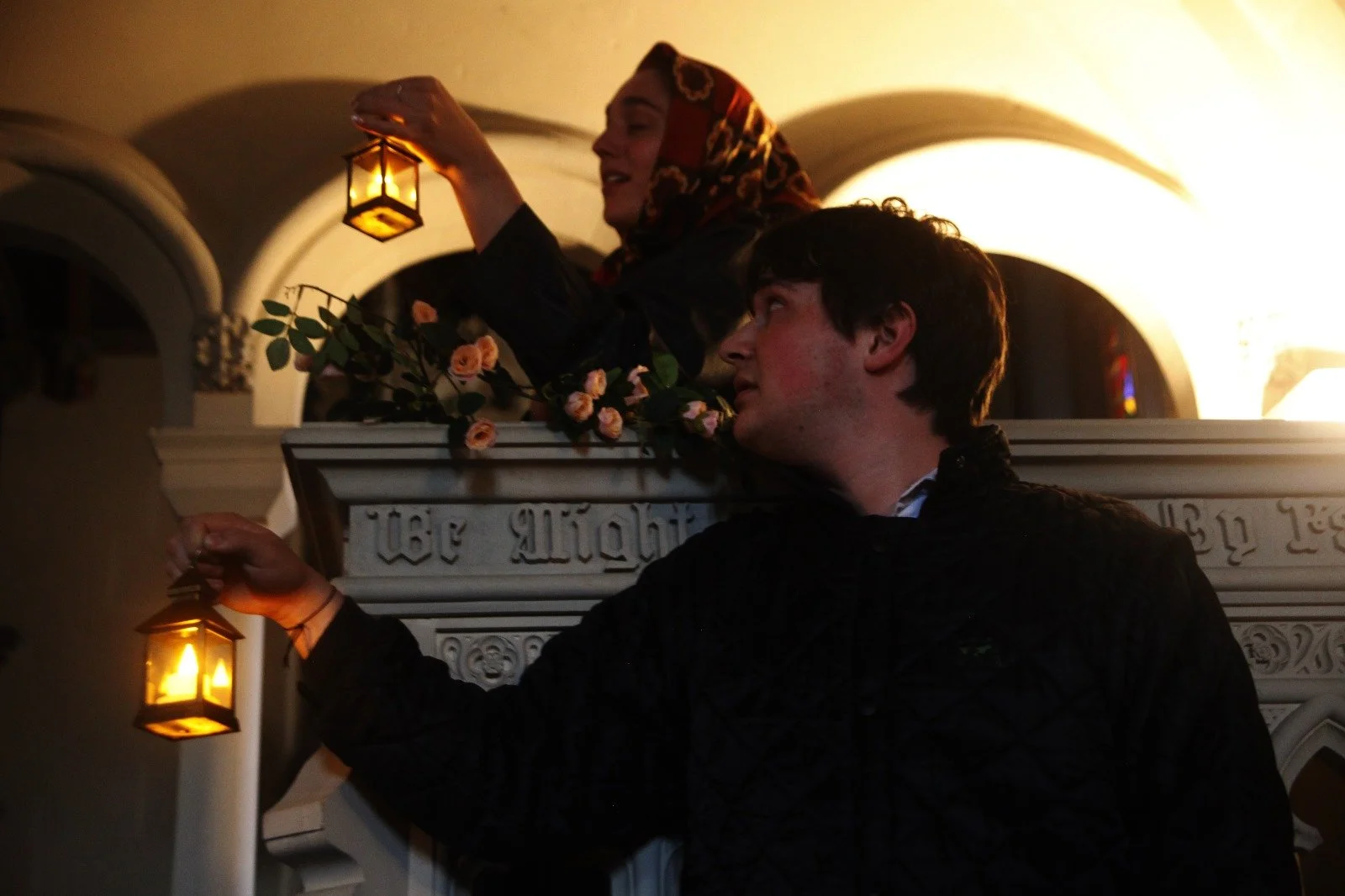 A woman in a headscarf holding a lantern while standing on a balcony, and a young man holding a lantern standing below the balcony with his arm outstretched. The setting appears to be a church or a religious building with decorative elements and warm
