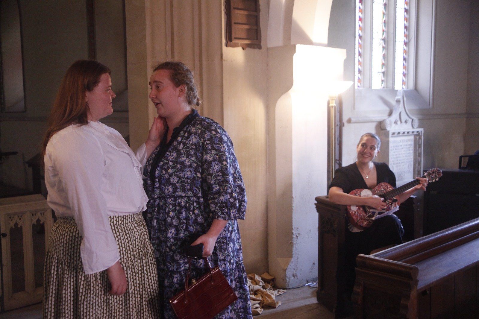 Two women are engaging in conversation inside a church or chapel, one with long brown hair in white blouse and patterned skirt, the other with blonde hair in a floral dress holding a phone and purse. A woman is sitting in the background playing a gui