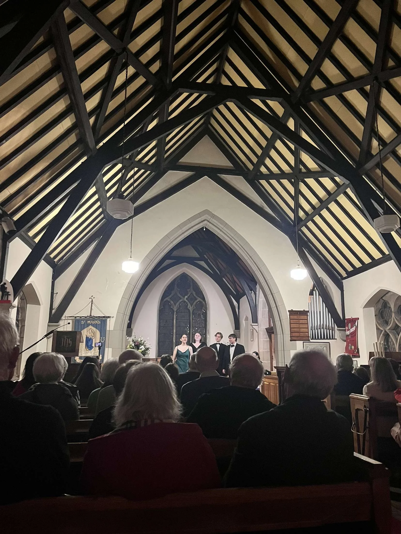 People singing in a church during a performance, with an audience sitting in pews, stained glass windows, and church banners visible.